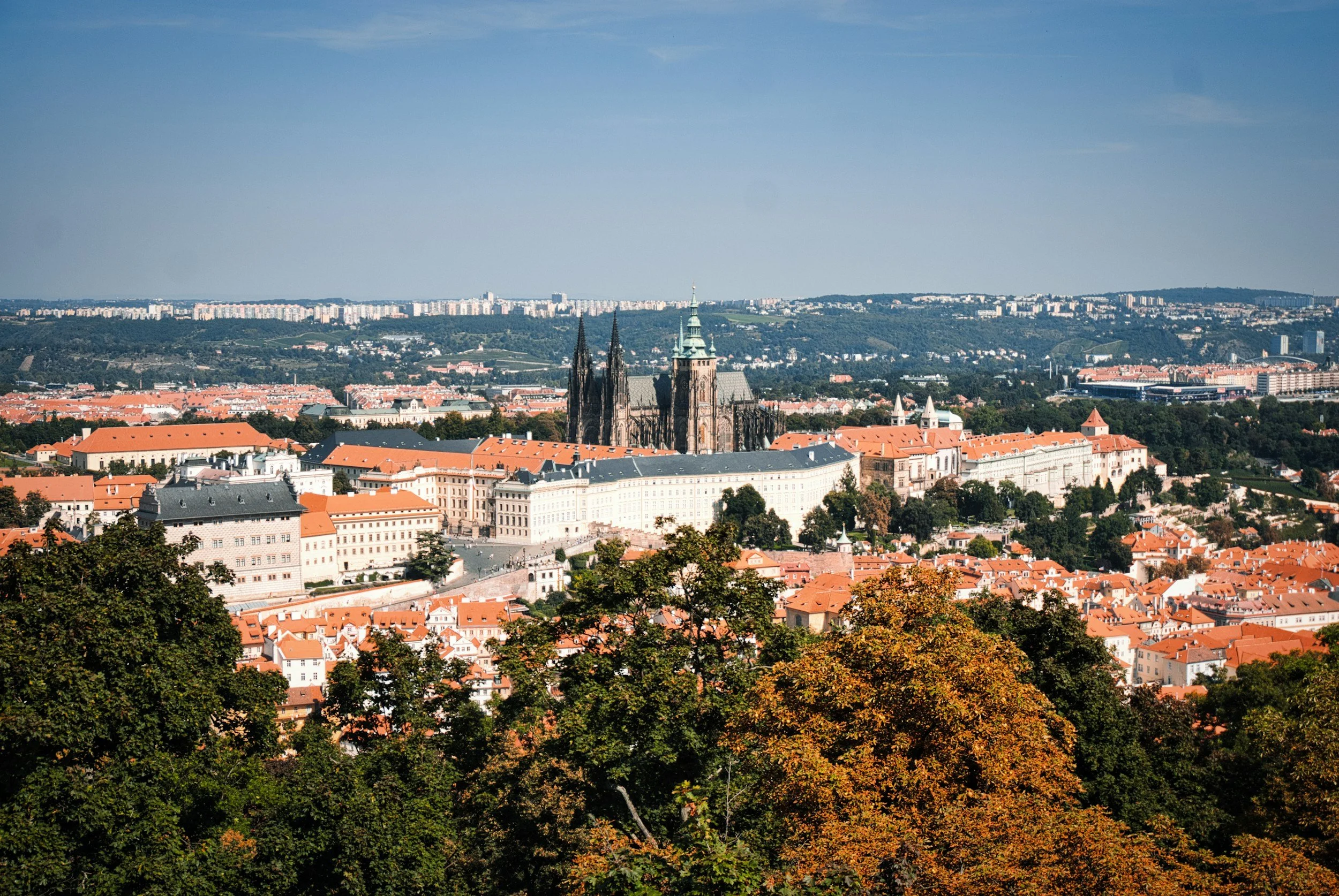 A panoramic view of a city skyline featuring a prominent Gothic cathedral with tall spires, surrounded by historic buildings with orange rooftops, and trees with green and autumn-colored leaves in the foreground.