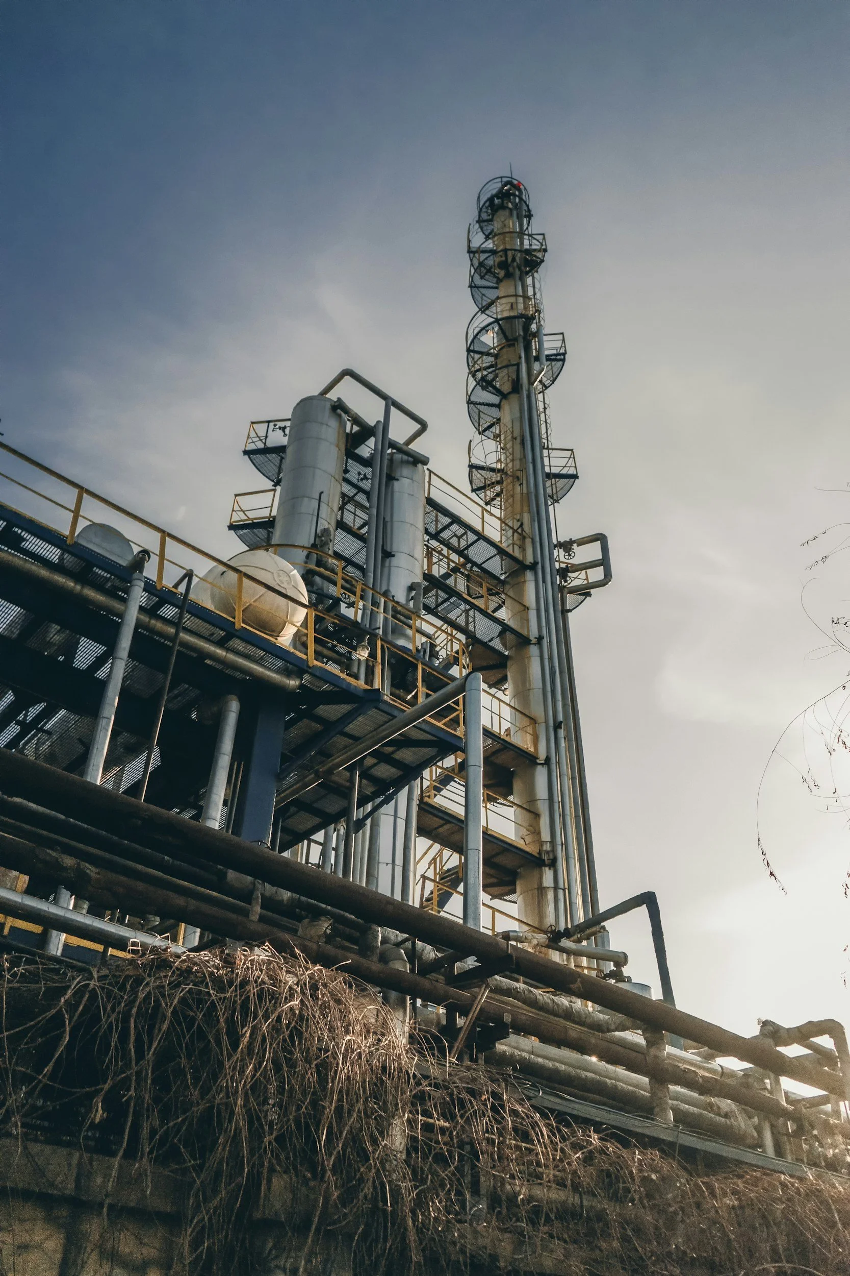 Industrial refinery with tall distillation tower, pipes, and staircases, against a cloudy sky.