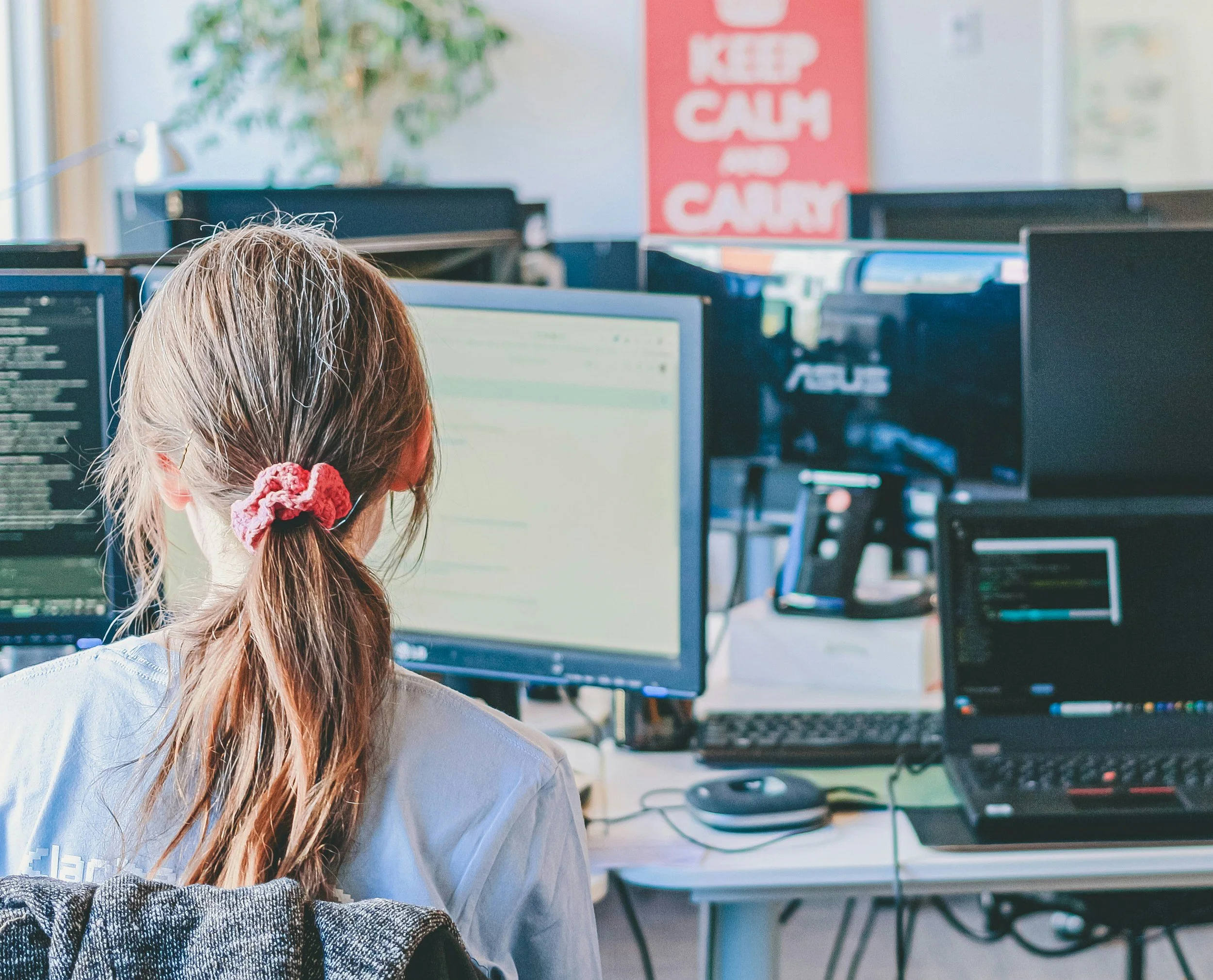 A woman with light brown hair tied back with a pink scrunchie, sitting at a desk in front of multiple computer monitors in an office. A red poster on the wall reads 'KEEP CALM AND CARRY'.