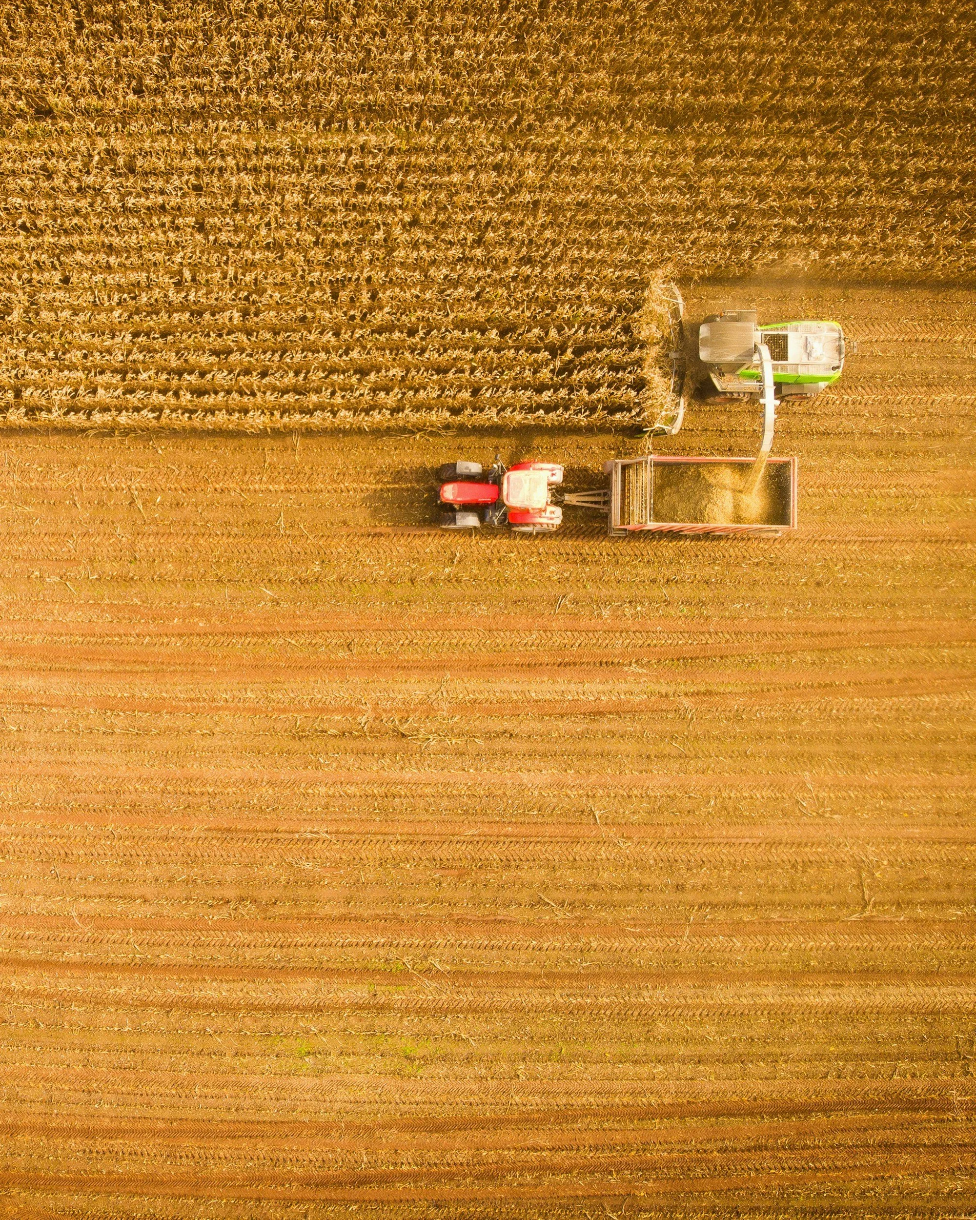 An aerial view of a farm field with a red tractor and a green harvester, harvesting crops in a large, open field.