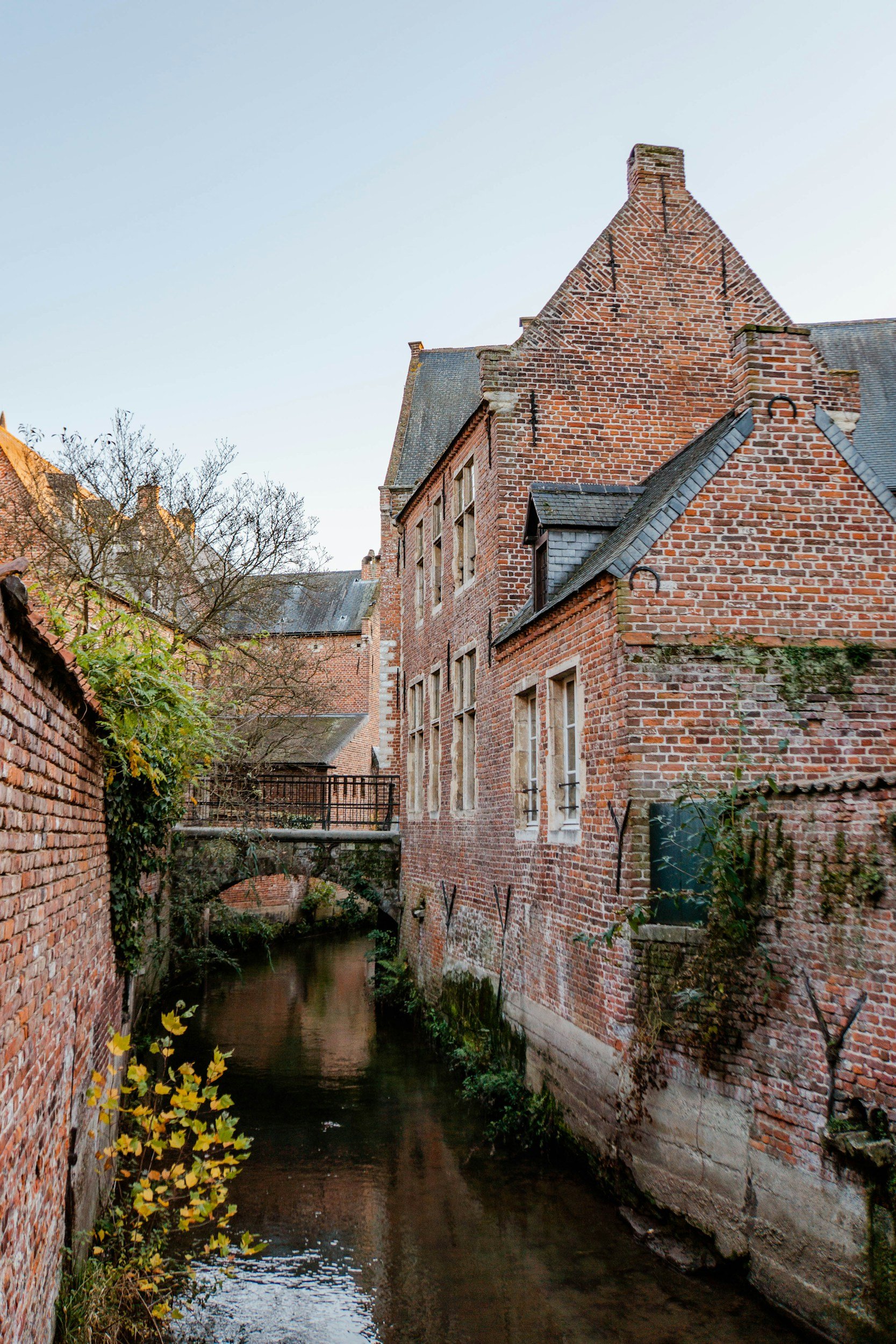 Old brick buildings along a narrow canal with a small stone bridge, in a historic European town.
