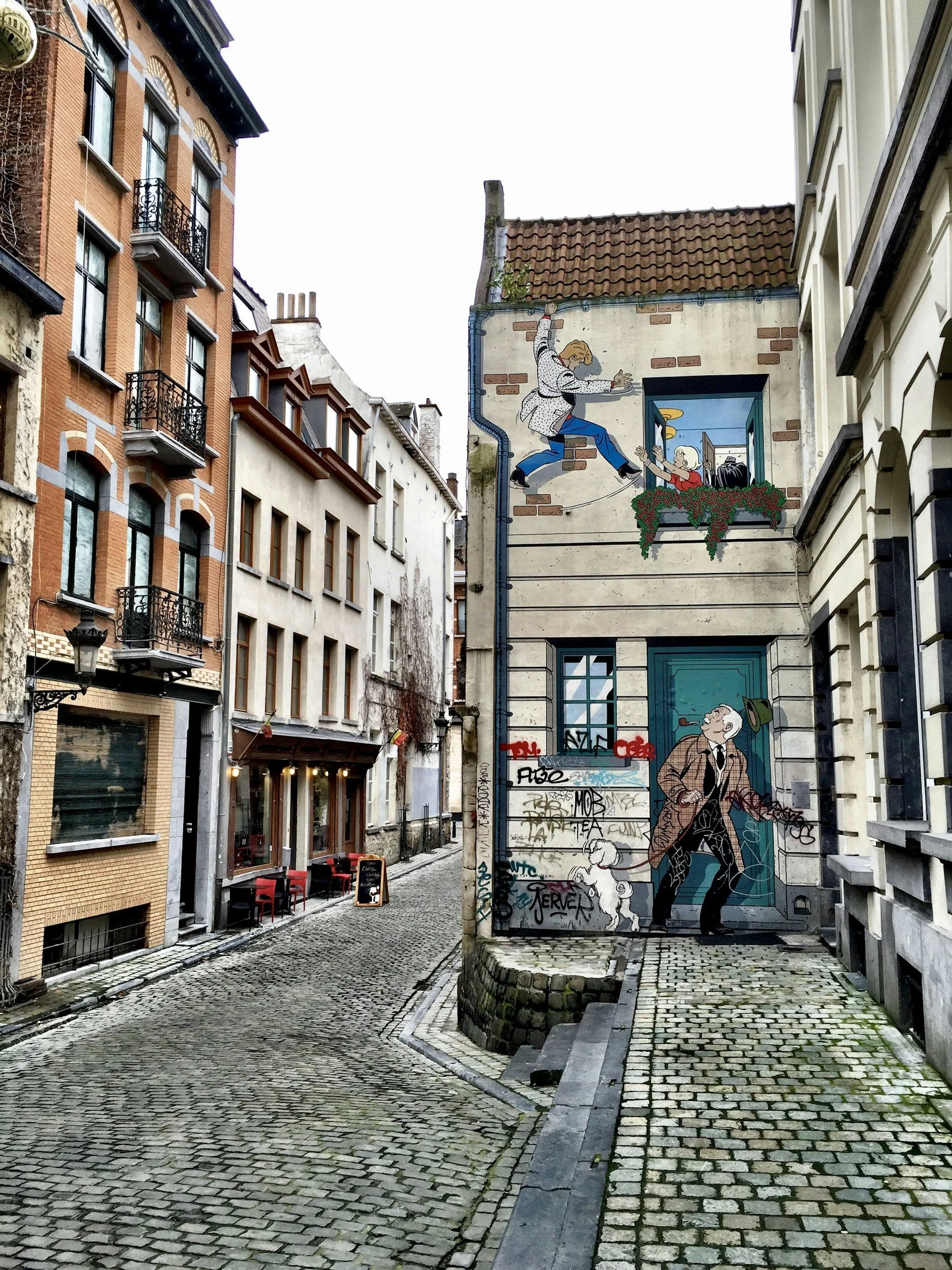 Street scene with cobblestone alley, buildings on either side, and a mural on a building featuring characters from Tintin. The mural depicts Tintin and Captain Haddock outside a window and a man walking a dog.