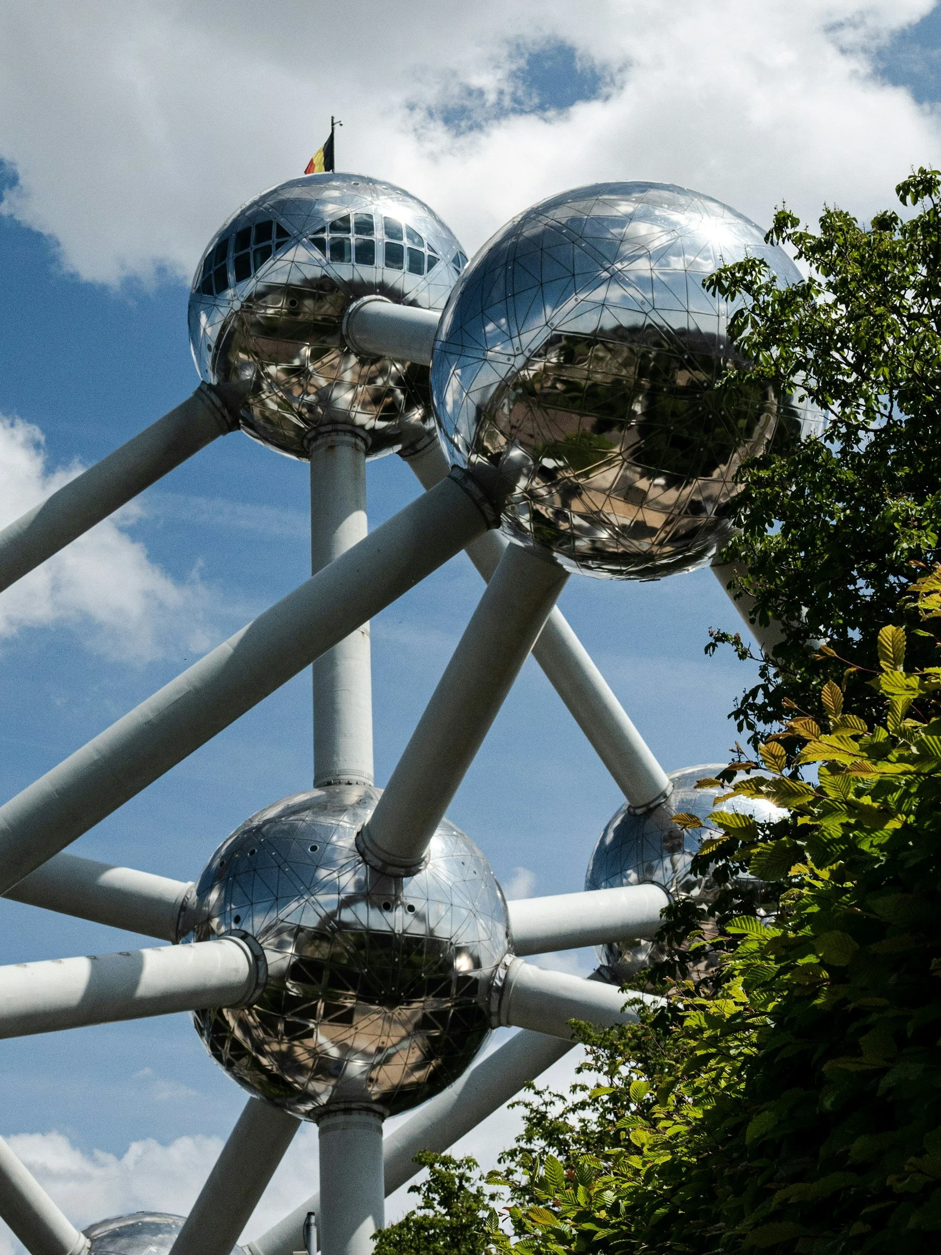 Close-up view of the Atomium structure in Brussels, Belgium, featuring large reflective metal spheres connected by white tubular beams with some green foliage in the foreground and a partly cloudy sky in the background.