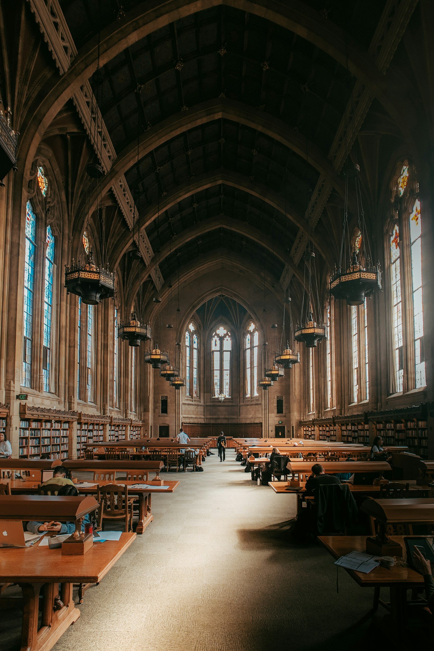 Interior of a large, historic library with high vaulted ceiling, tall stained glass windows, wooden bookshelves, and students studying at long wooden tables.