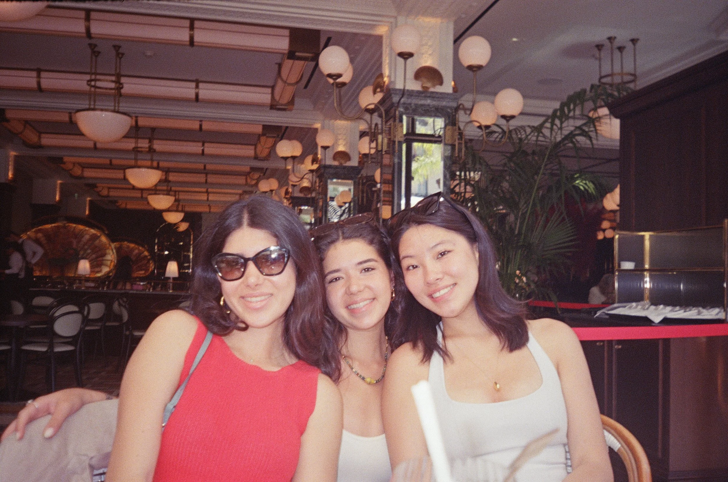 Three women smiling at a restaurant, one wearing sunglasses and a red sleeveless top, one in a white top with necklace, and the third in a white sleeveless top with sunglasses on her head, sitting together at a table.