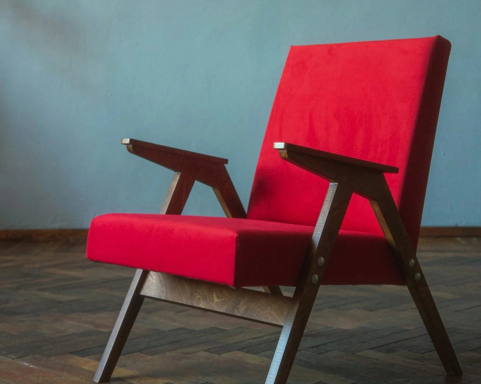 A mid-century modern armchair with a red upholstered seat and backrest, wooden armrests, and angled wooden legs, placed on a wooden floor against a light blue wall.