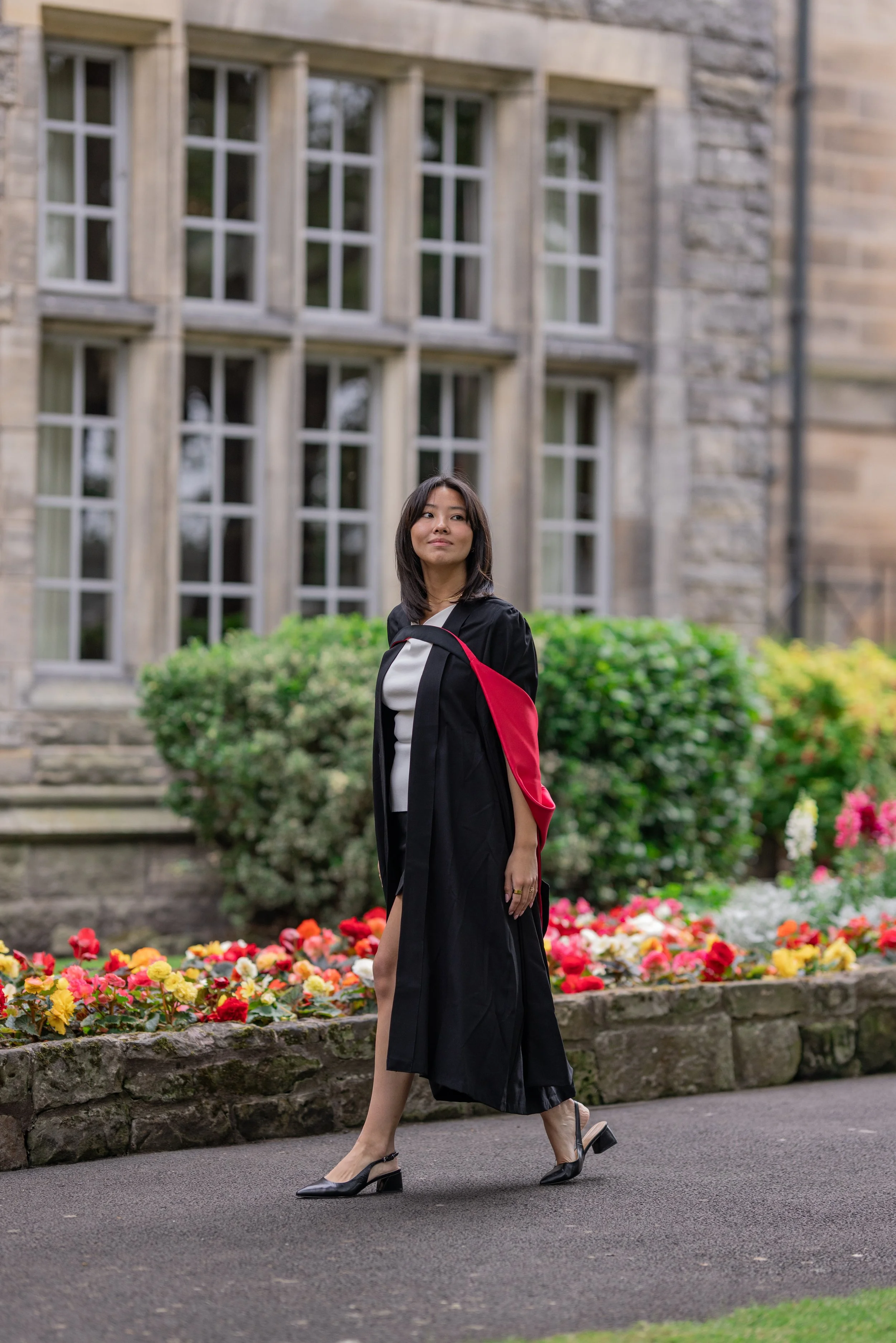 A woman in a graduation gown and heels walking on a pathway with colorful flowers and a large stone building with many windows in the background.