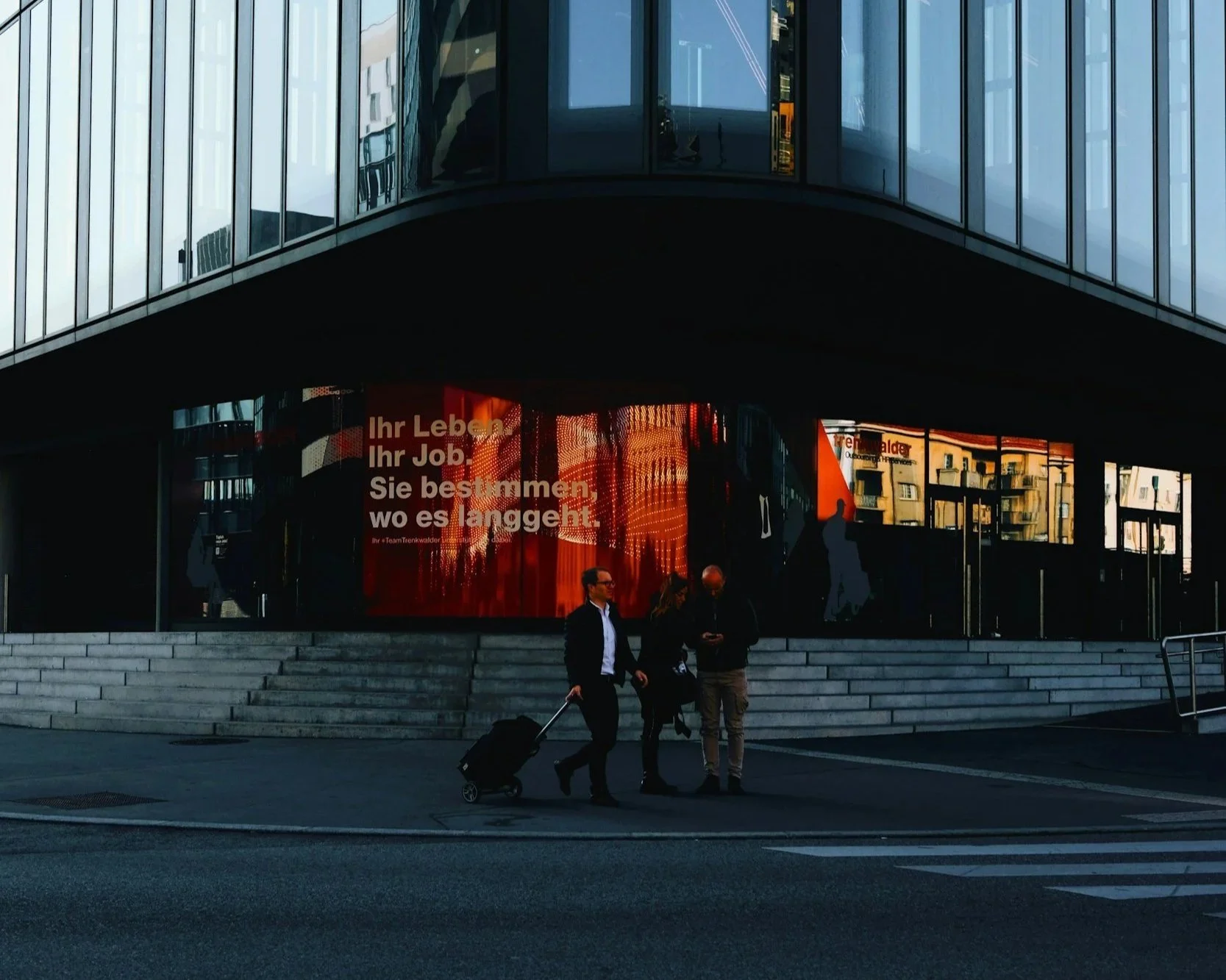 People walking past a modern building with a digital display showing German text.