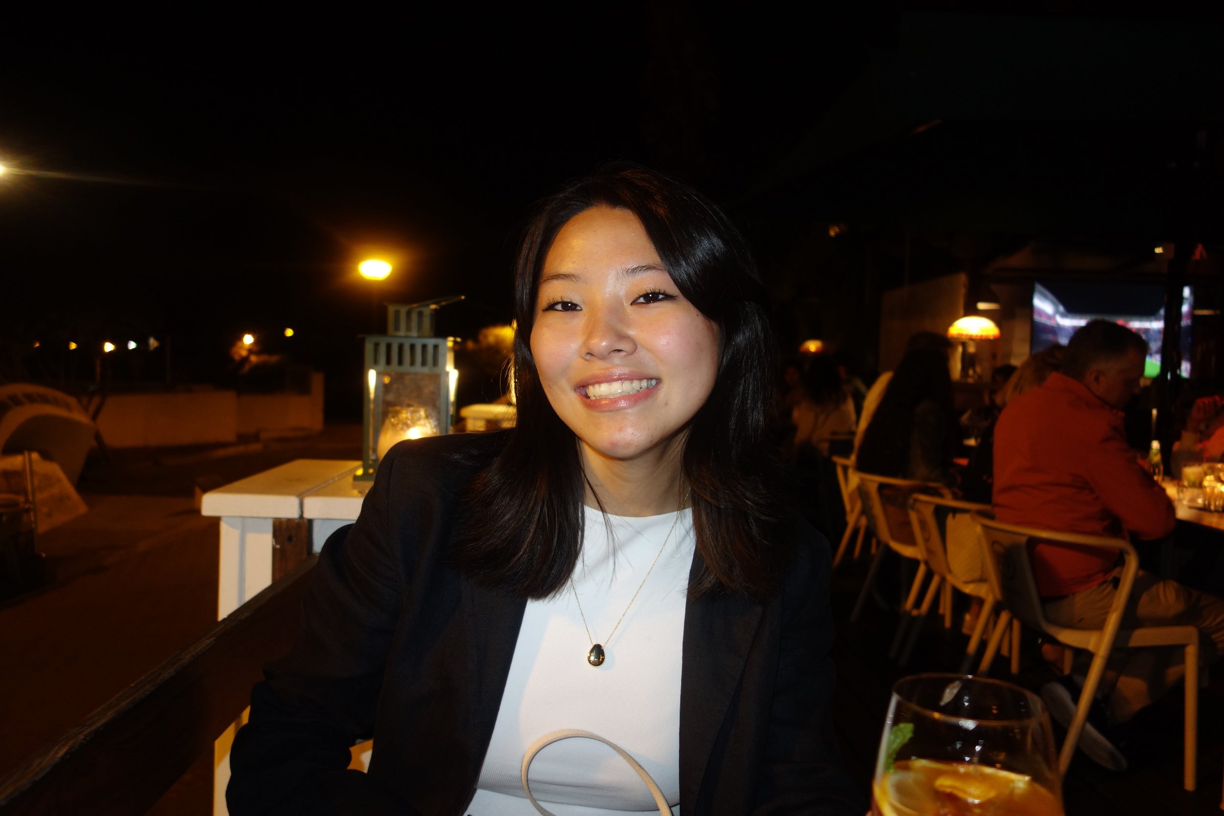 A young woman with shoulder-length black hair, smiling, wearing a black blazer and a white top, sitting at an outdoor restaurant at night with warm lighting and other diners in the background.
