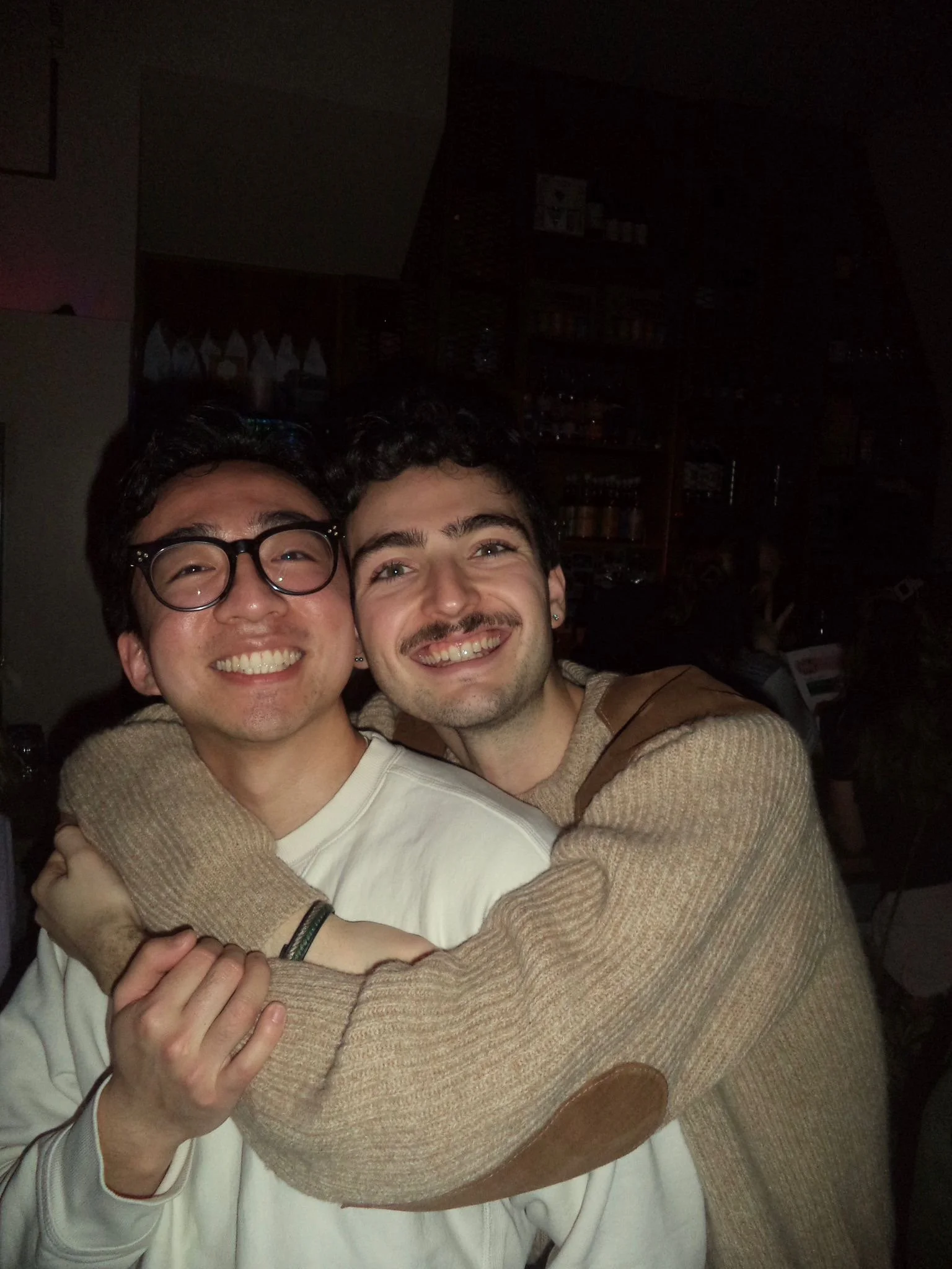 Two young men smiling and hugging each other indoors in a dimly lit room.