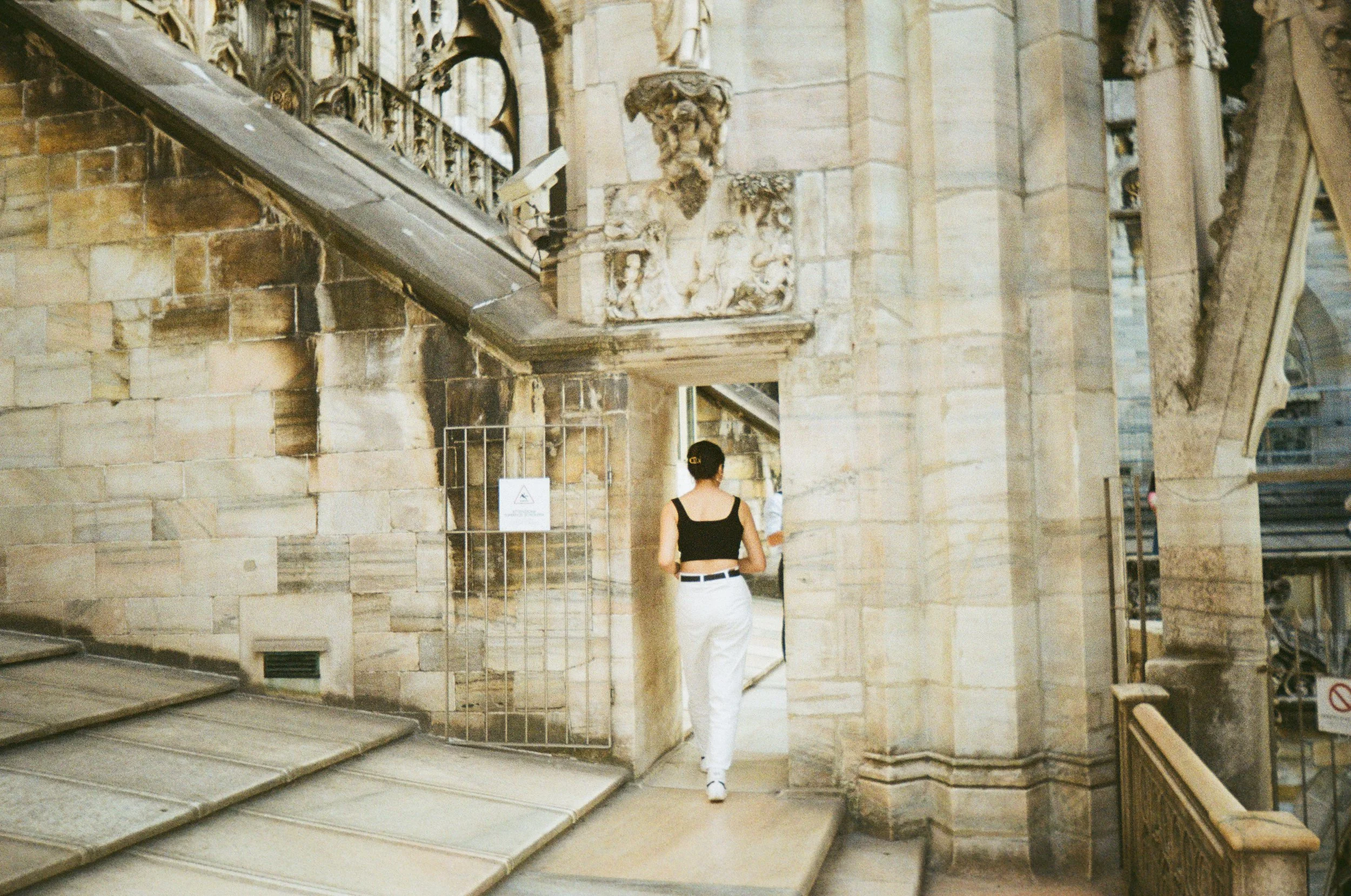A woman with dark hair in a bun, wearing a black sleeveless top and white pants, walking through a stone archway in a historic building with ornate architectural details.
