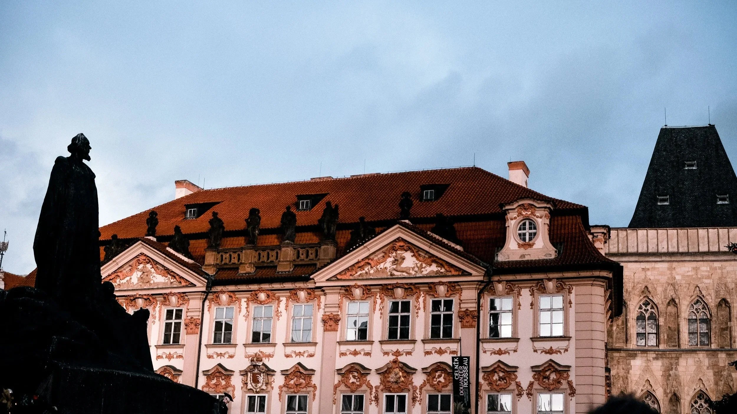 An ornate historic building with decorative details on its pink facade, a red-tiled roof, and a series of statues along the roofline. In the foreground, there's a silhouette of a statue of a person on a pedestal. The sky above is cloudy.