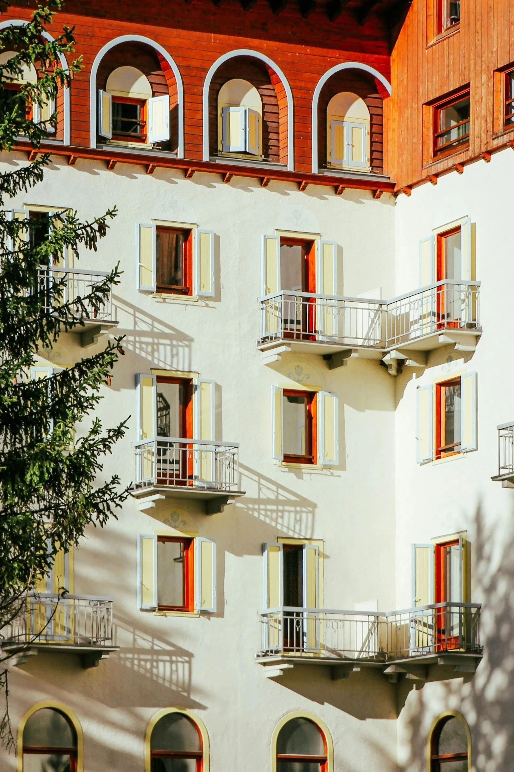 A multi-story building with white and wooden exterior, multiple open windows with shutters, small balconies, and a shadow of a tree cast on the wall.