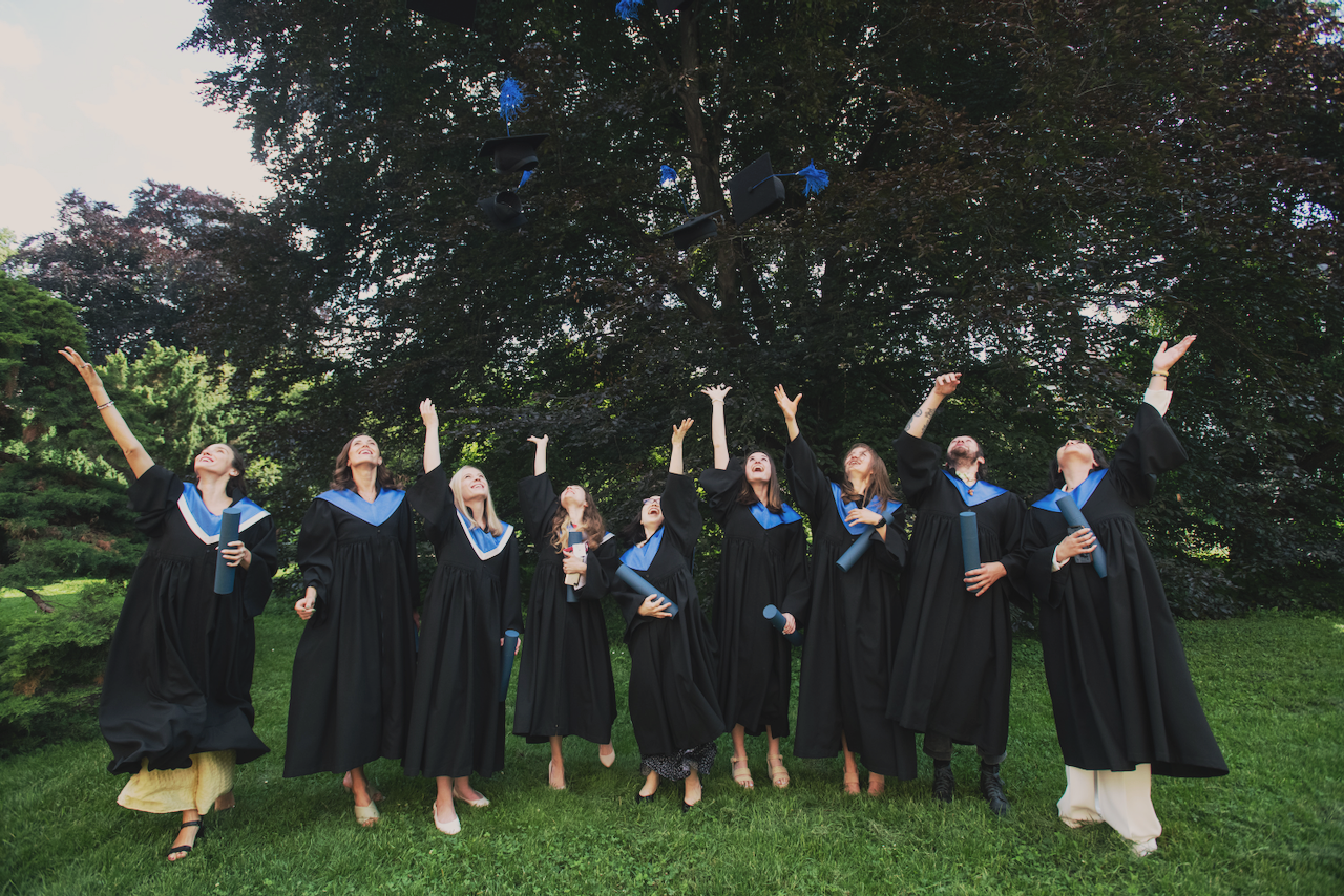 Group of graduating students in caps and gowns celebrating outdoors on a grassy field, tossing their caps into the air.