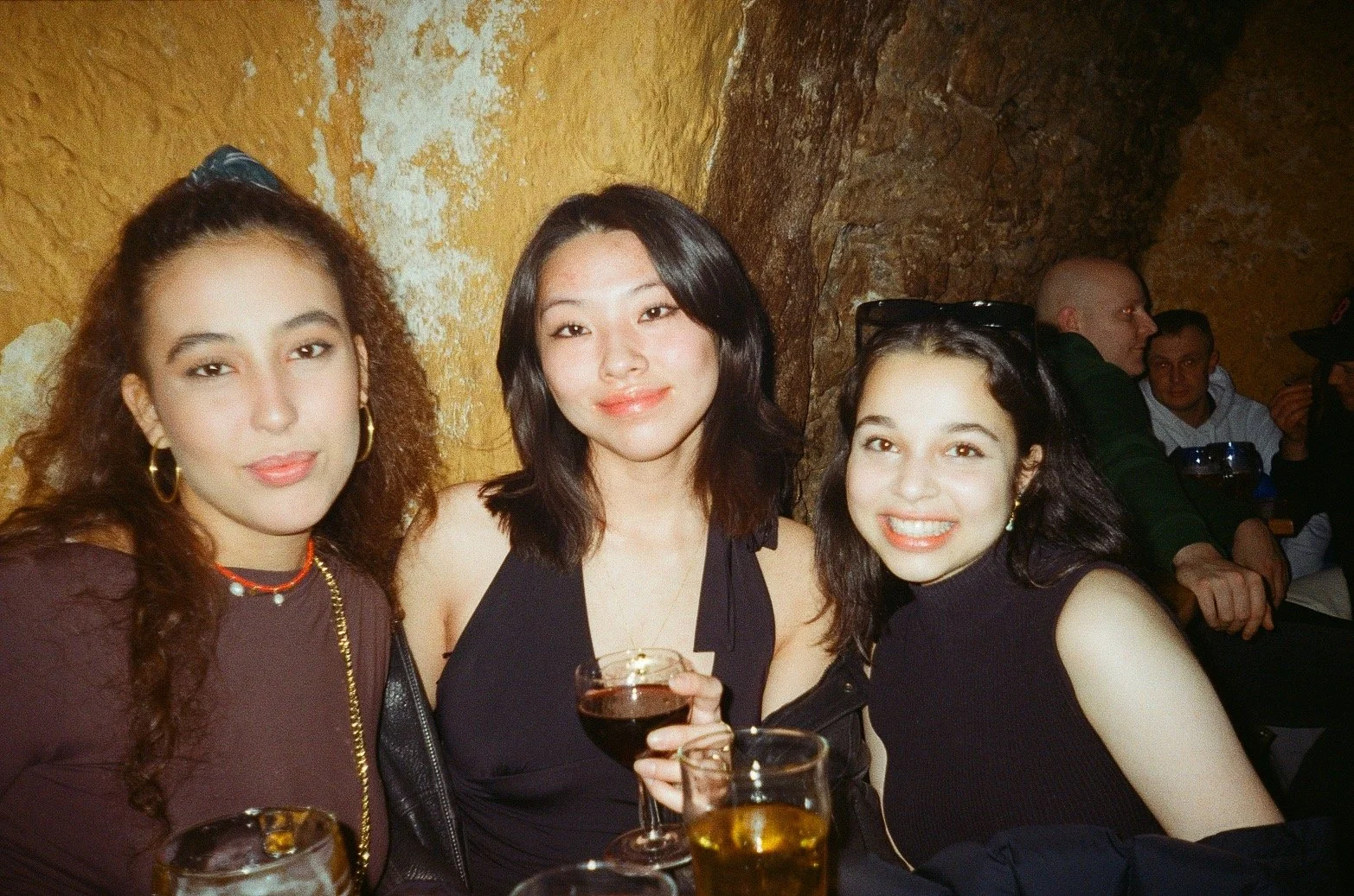 Three women sitting together at a bar or restaurant, smiling for the camera, with drinks in front of them. They are dressed in fashionable clothes and the background shows a rustic, cave-like wall with other people sitting at tables.