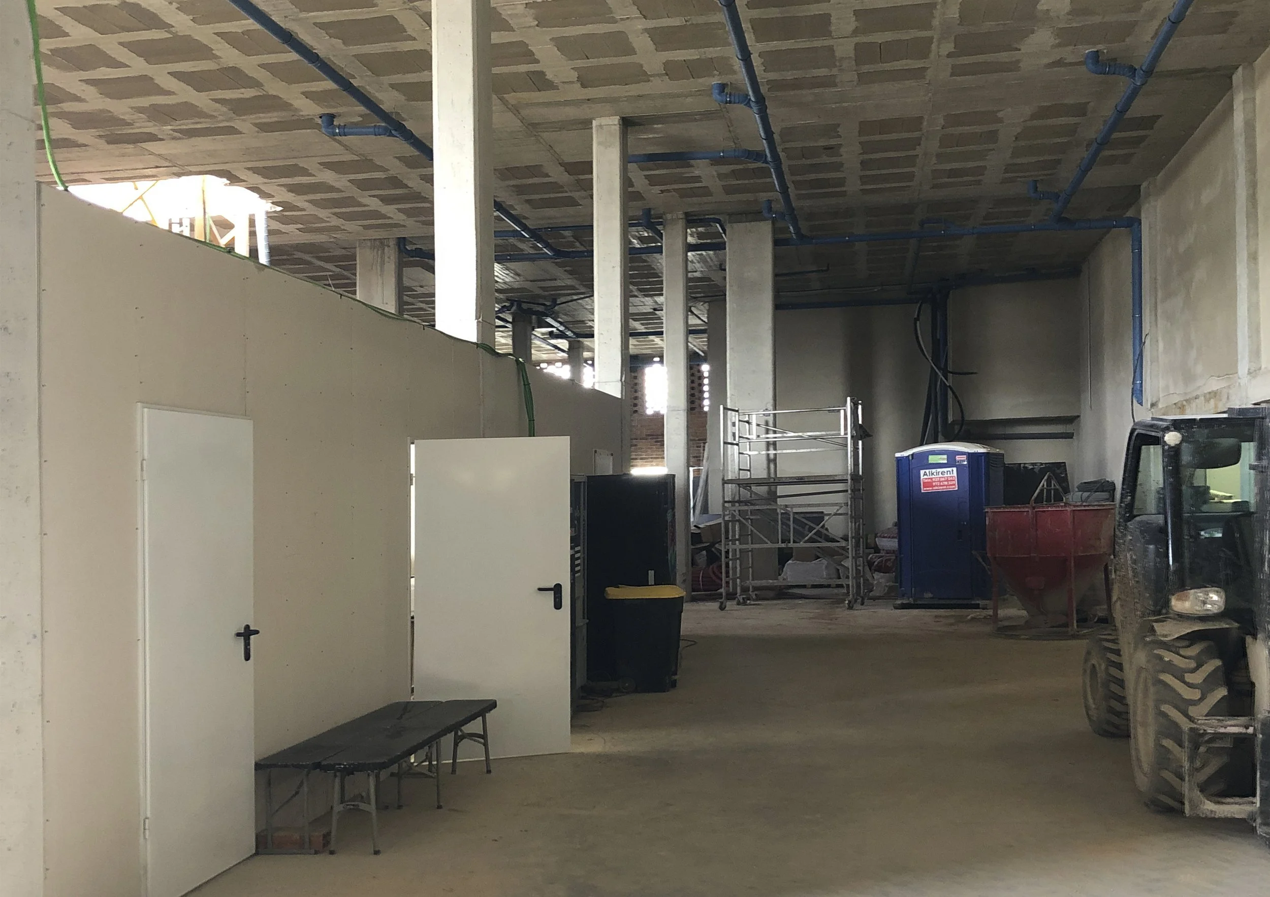 Espacio de construcción interior con columnas de concreto, tuberías azules en el techo, maquinaria y equipo de construcción, y una pared blanca con una puerta y un banco en primer plano.