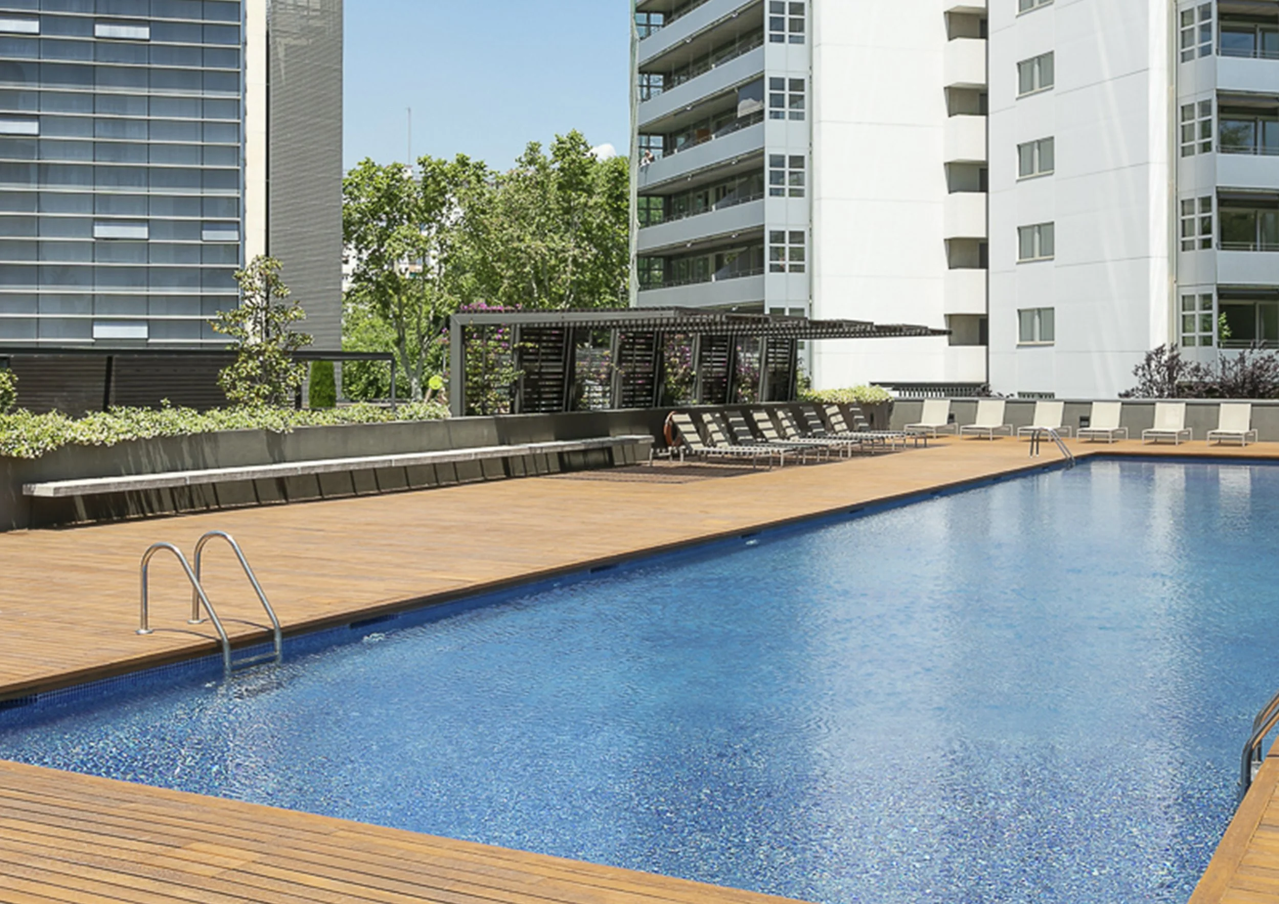 Área de piscina en un complejo residencial con sillas y mesas, edificios altos y vegetación en el fondo.