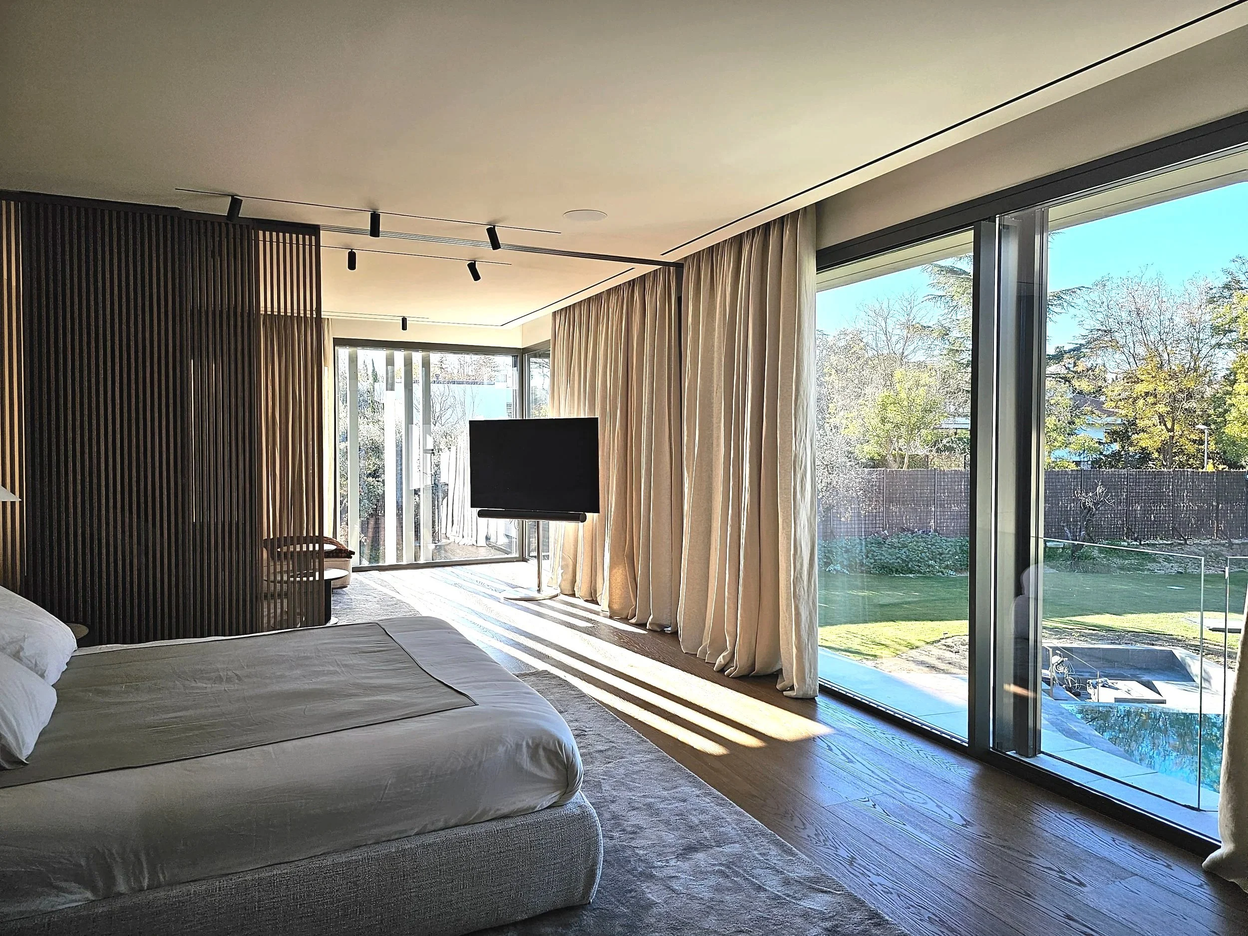Interior de una habitación moderna con cama, cortinas claras, paredes de madera y ventanas grandes que dejan ver un jardín con árboles y césped.