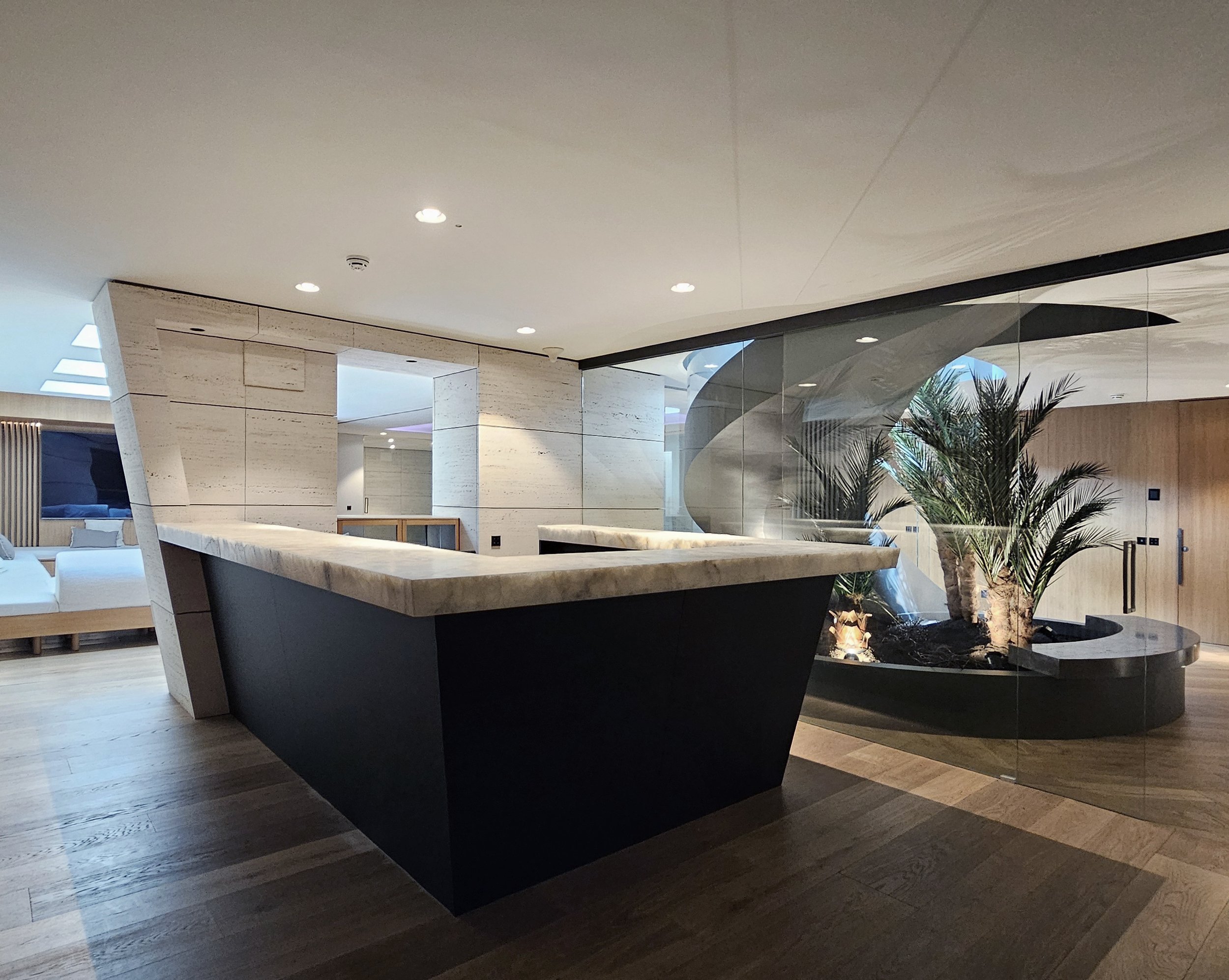 Interior moderno con mobiliario de madera clara, área de descanso con sofá blanco y plantas decorativas, iluminación empotrada y paredes de piedra y madera.
