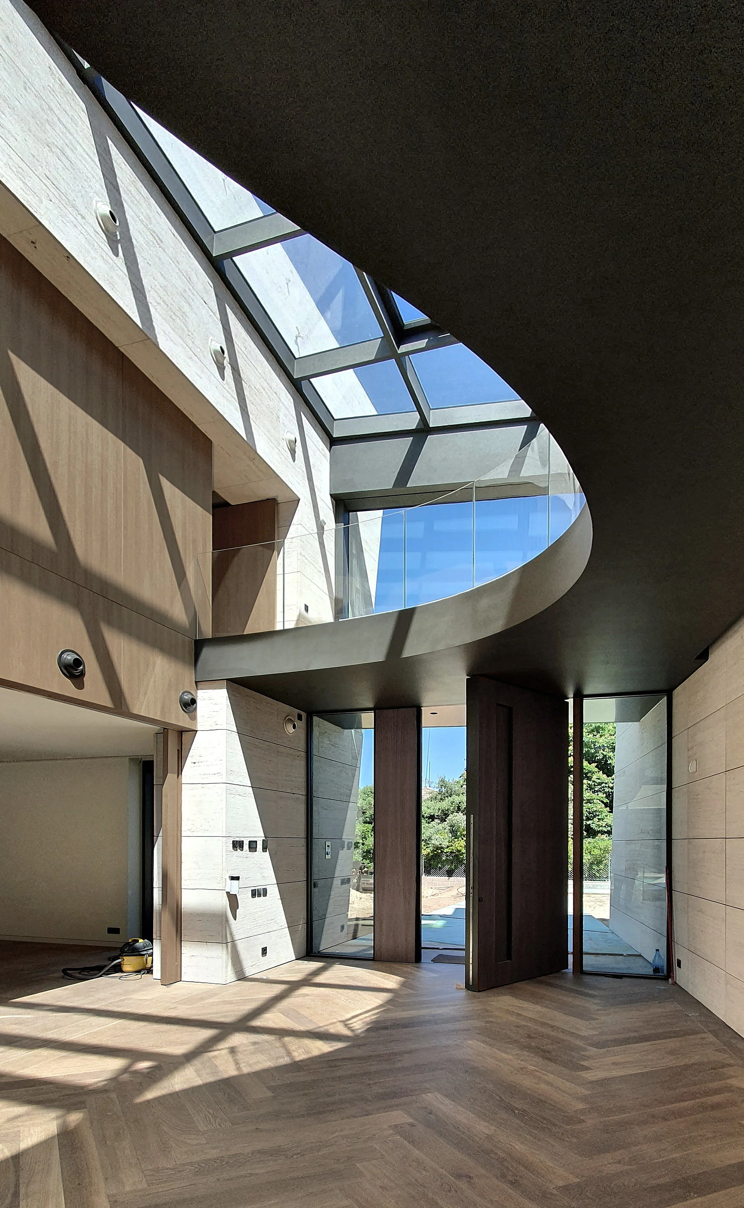 Interior de una casa moderna con paredes de piedra, doble altura, ventanales de vidrio y madera, y piso de madera en patrón diagonal.