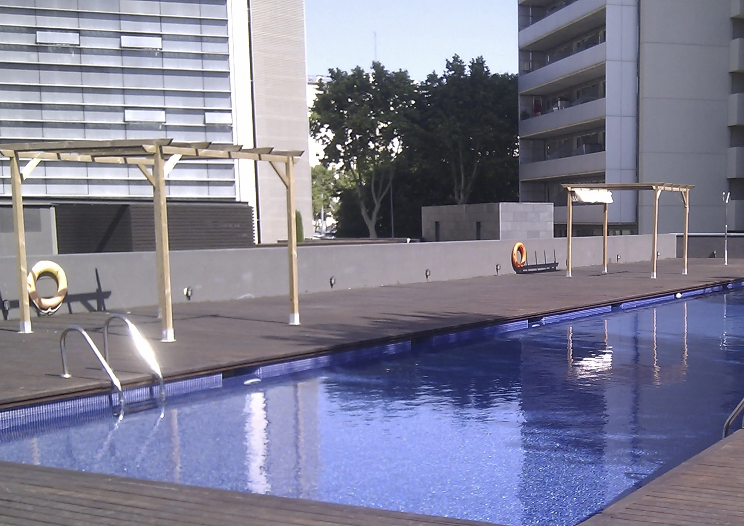 Piscina en un edificio moderno, con estructuras de madera junto a la piscina y edificios de apartamentos con árboles al fondo