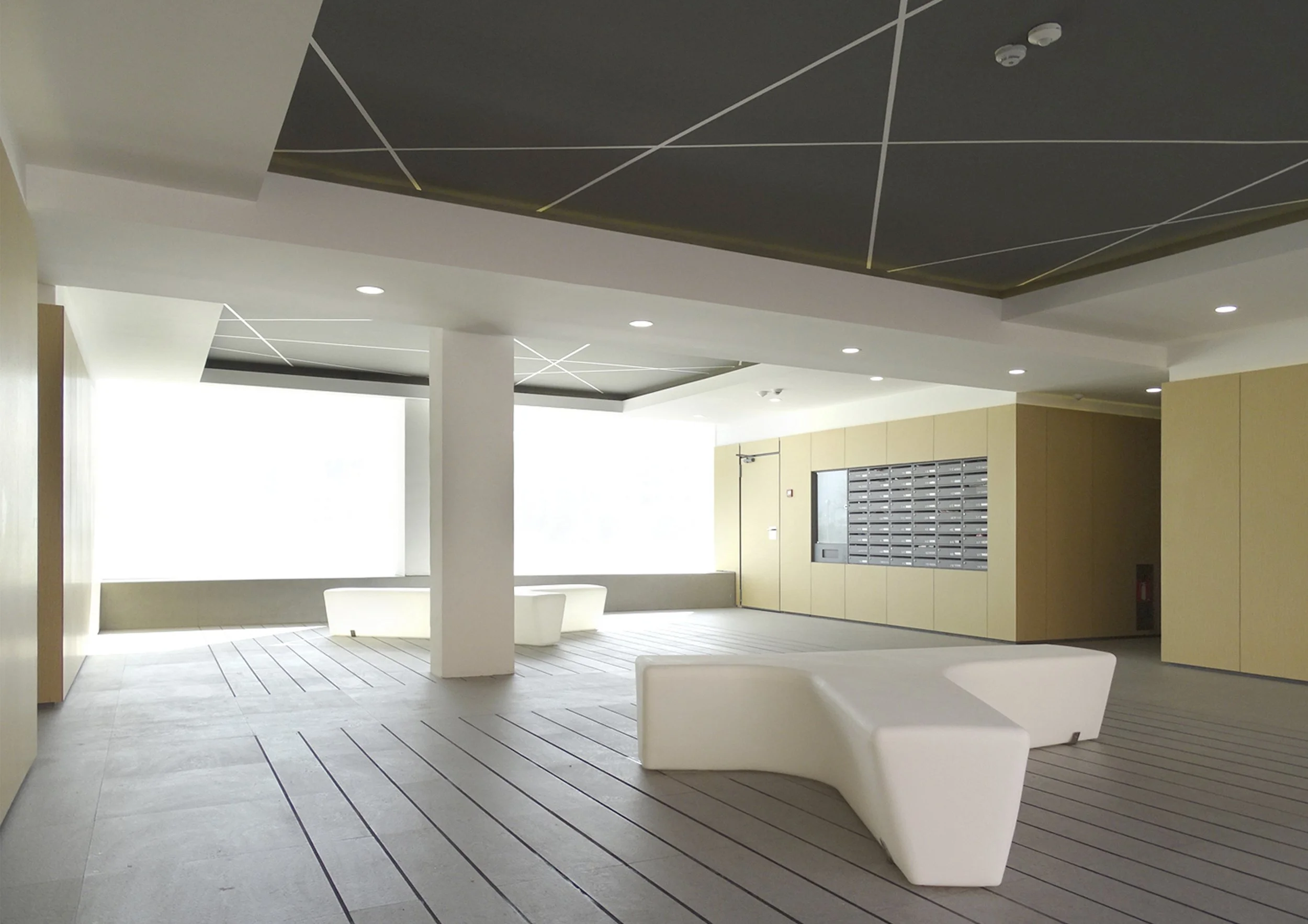 Interior moderno de un edificio con paredes blancas, techo gris con líneas blancas, y mobiliario en tonos claros y oscuros. Hay bancos blancos en primer plano y una pared con buzones en el fondo.