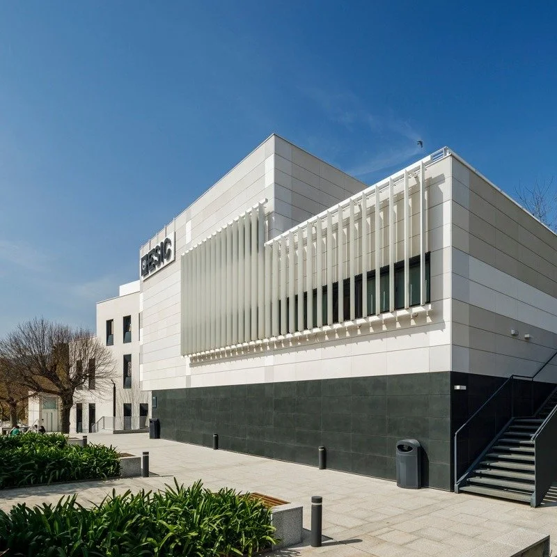 Edificio moderno con fachada blanca y ventanales con persianas verticales, área peatonal con árboles y zanjas de vegetación.