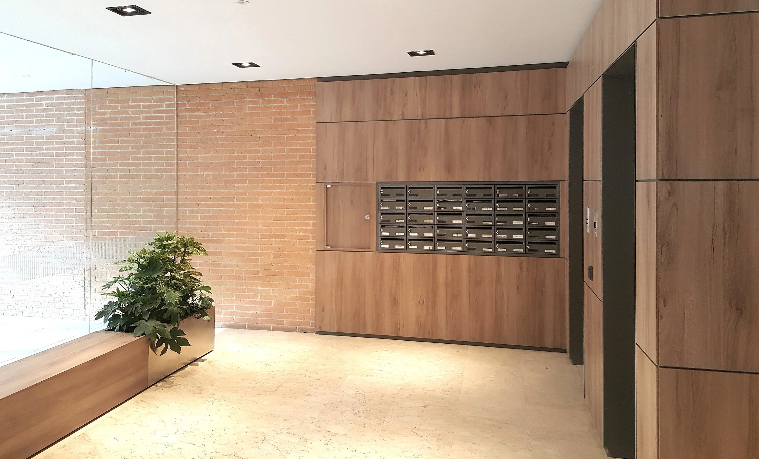 Interior de un lobby de edificio con paredes de ladrillo y madera, buzones de correo, planta decorativa y ventanas de vidrio.