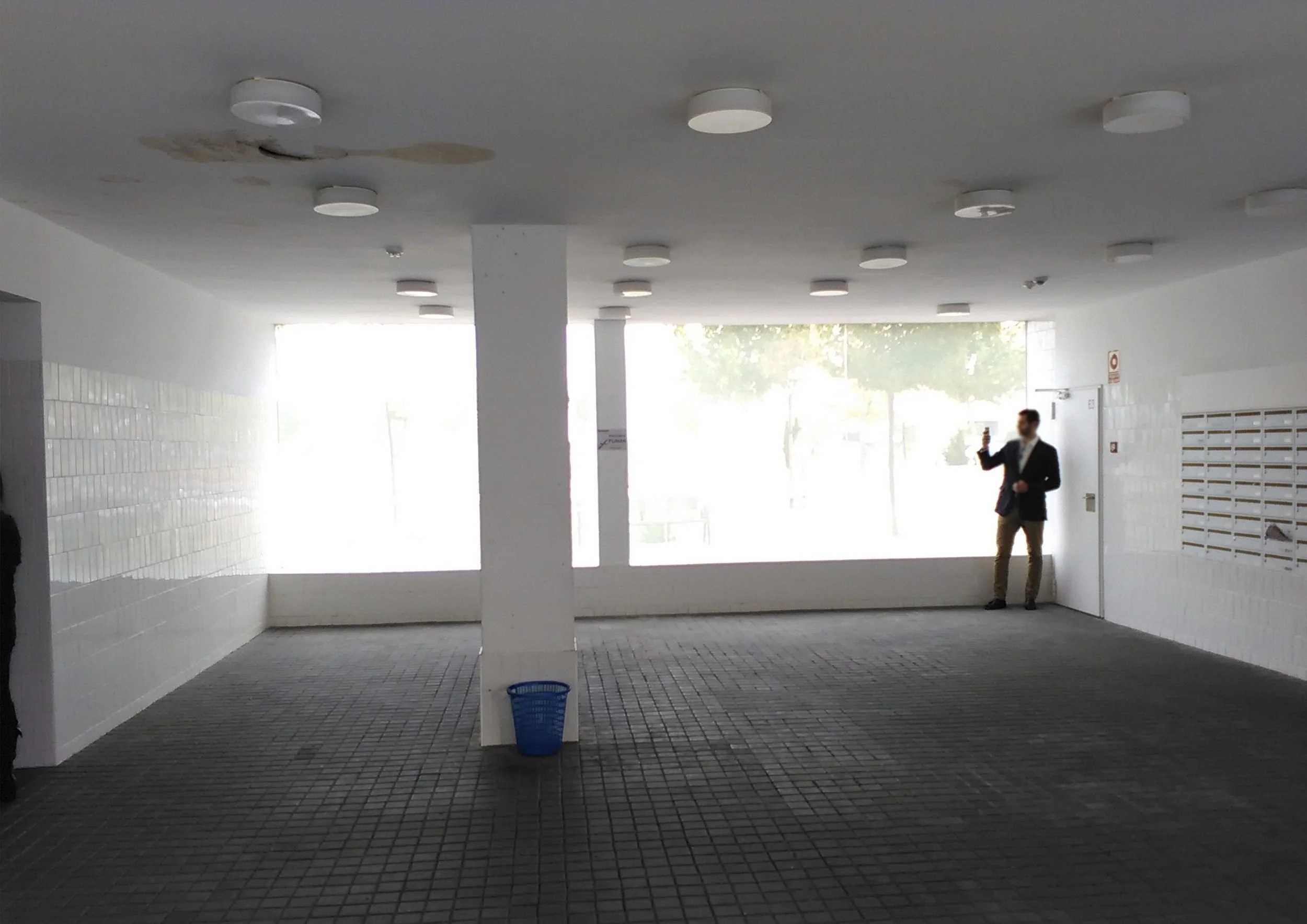 Pasillo cubierto con techo blanco, múltiples luces redondas en el techo, paredes blancas con azulejos en la parte inferior, suelo de mosaico, a la derecha hay cambios de casilleros metálicos y una señal de advertencia en la pared, en el fondo una mujer de traje con móvil en la mano, de cabello corto y oscuro, tomando una selfie o video.