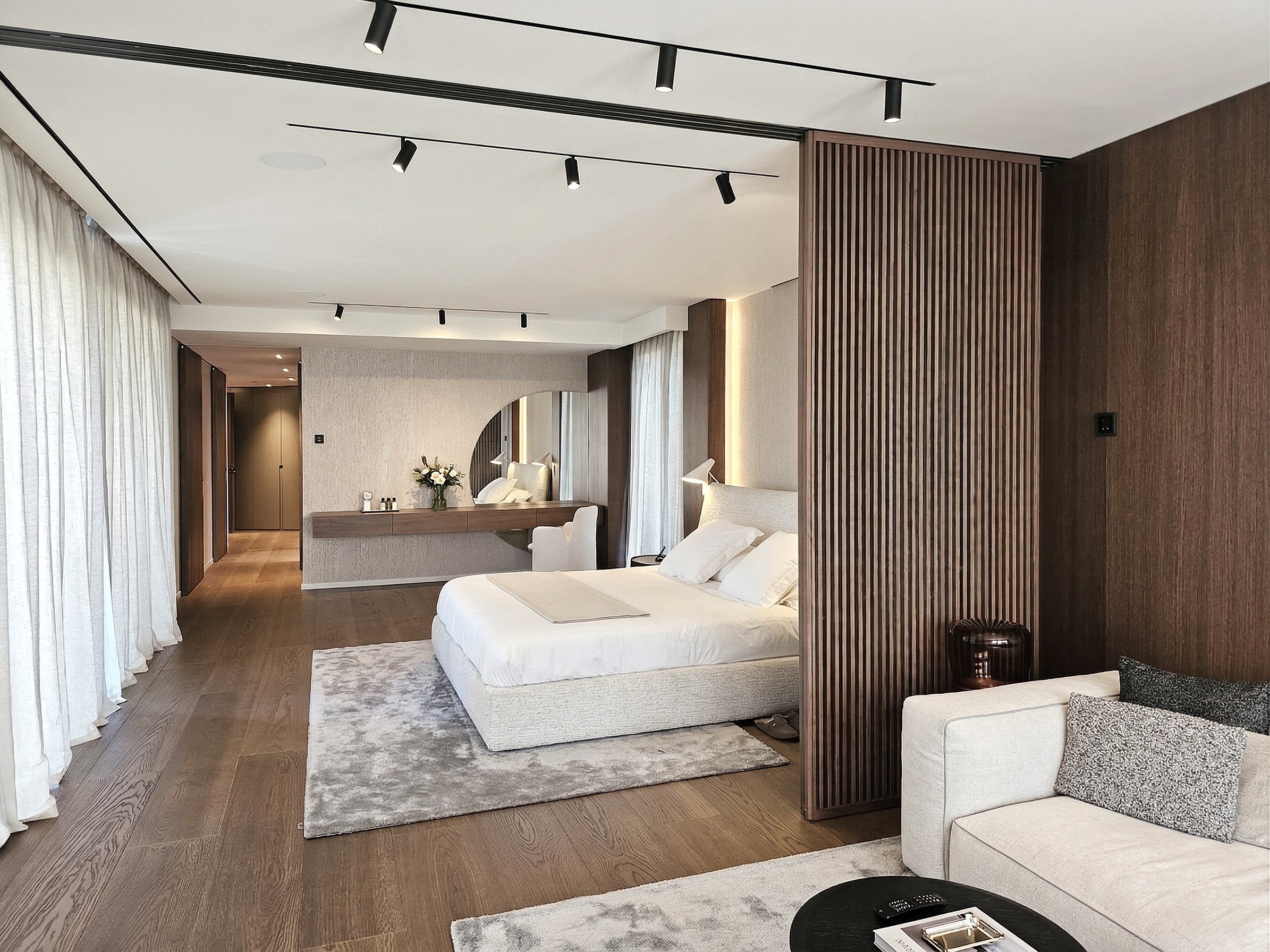 Habitación moderna con cama, sillón, mesa de noche, cortinas blancas, paredes de tonos neutros, piso de madera y accesorios decorativos.