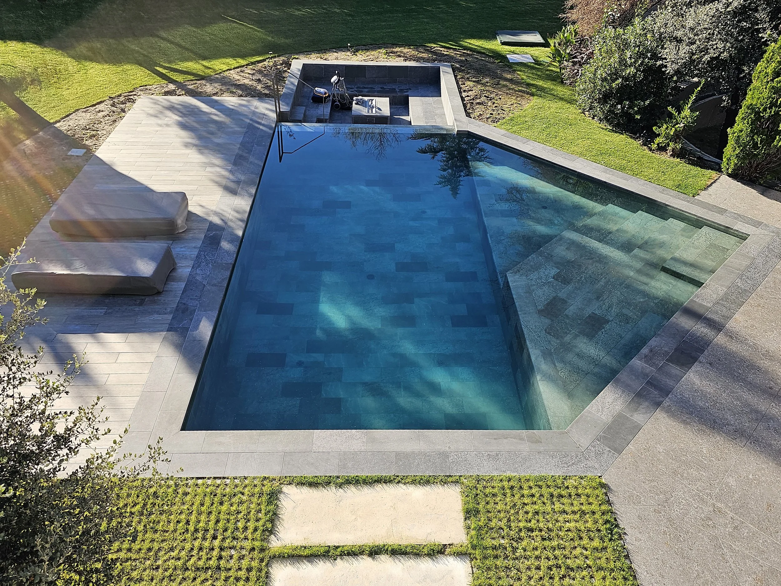 Piscina rectangular de agua azul clara en un patio con piso de piedra gris, rodeada de césped verde y árboles y arbustos en el jardín.