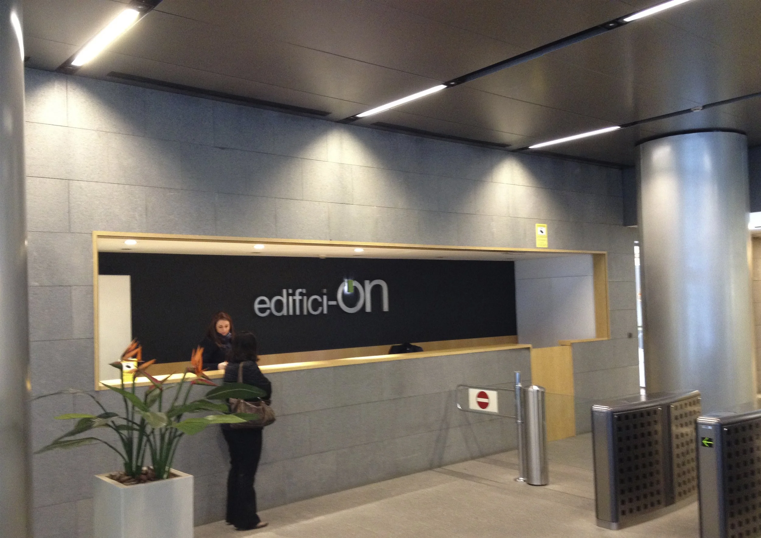 Mostrador de recepción en un edificio, con una mujer atendiendo a dos personas. Hay plantas en la esquina y una barra con un cartel que dice 'edifici-ON'. La pared es de color gris y el techo tiene iluminación moderna.