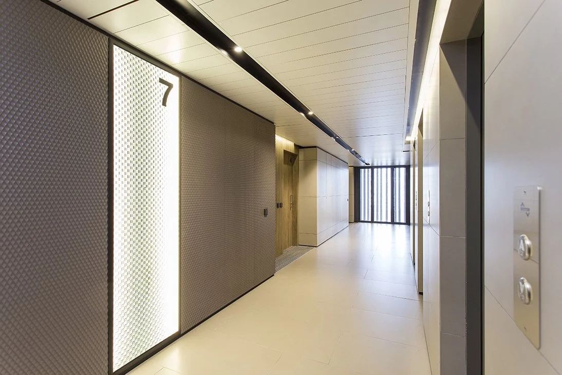 Pasillo de edificio moderno con elevador y puertas, iluminación suave, pisos claros y paredes de tonos neutros.
