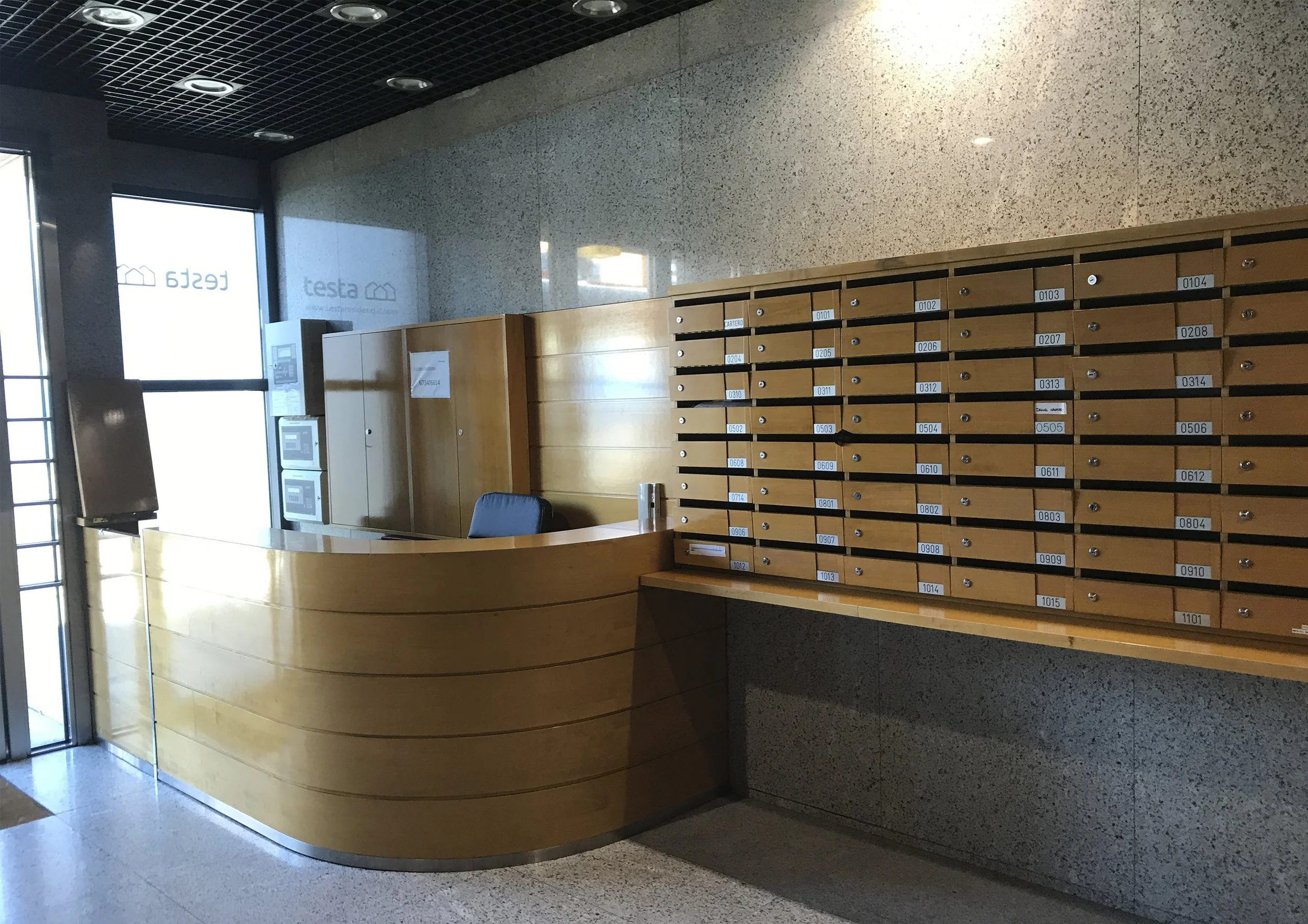 Mostrador de recepción en un edificio con buzones de madera y dos máquinas de pago, paredes de mármol y ventanas de vidrio.