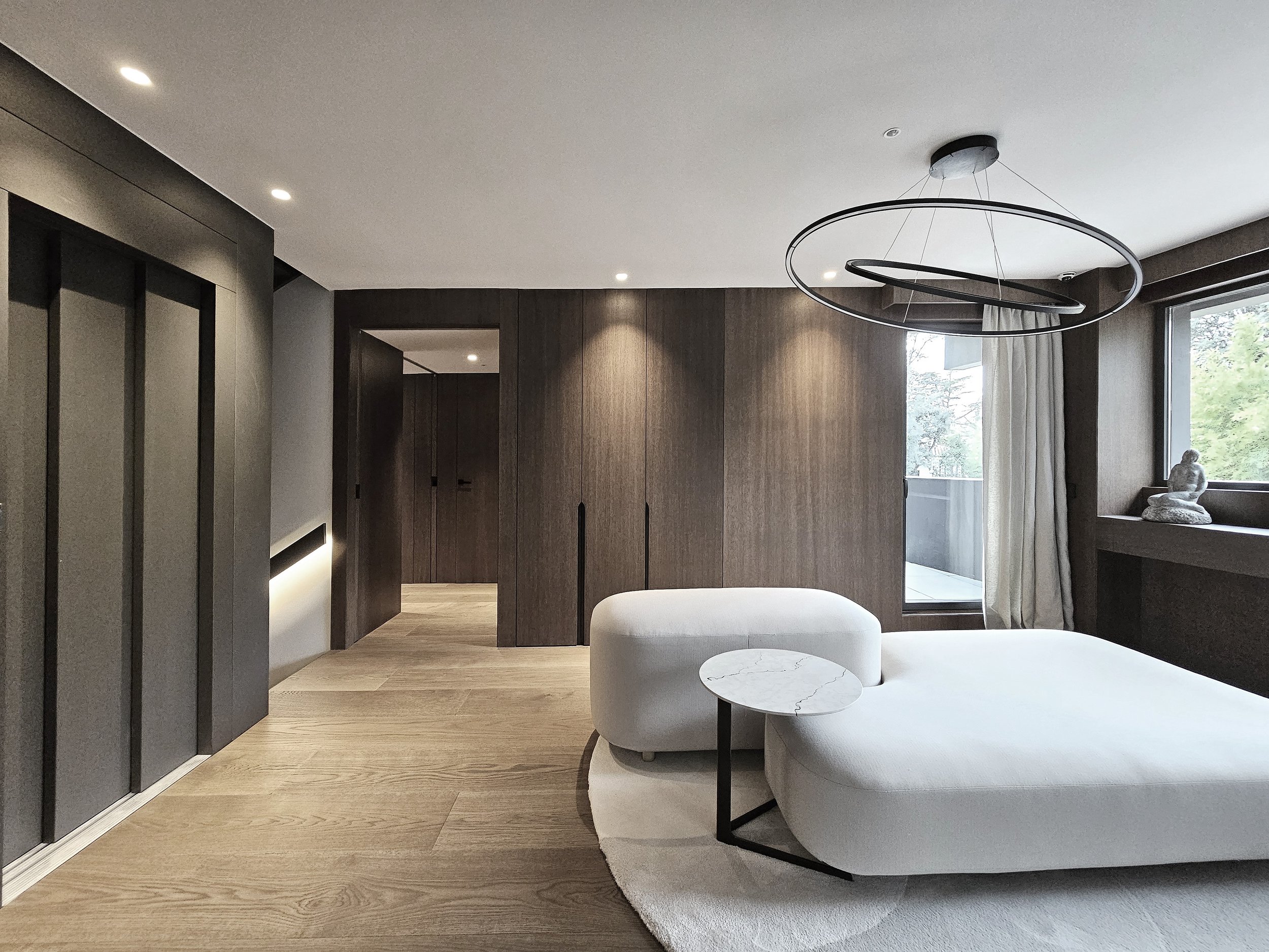 Interior moderno con paredes de madera, iluminación empotrada en techo, muebles blancos redondeados, mesa de mármol, grande ventana con cortinas blancas, figura de Buda decorativa en ventana.