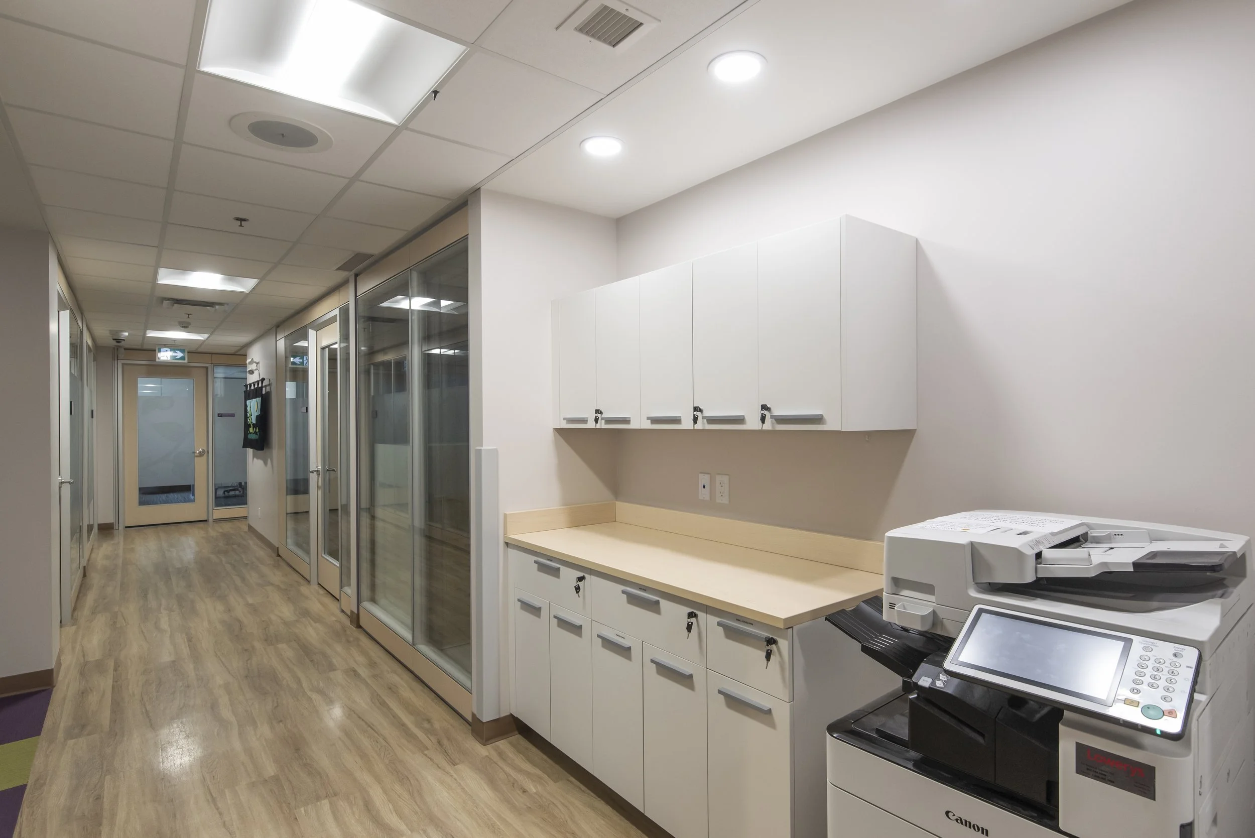 A modern office hallway with wooden flooring, glass enclosed meeting rooms on the left, and a small kitchenette area with white cabinets and a countertop on the right, including office equipment like a copier.