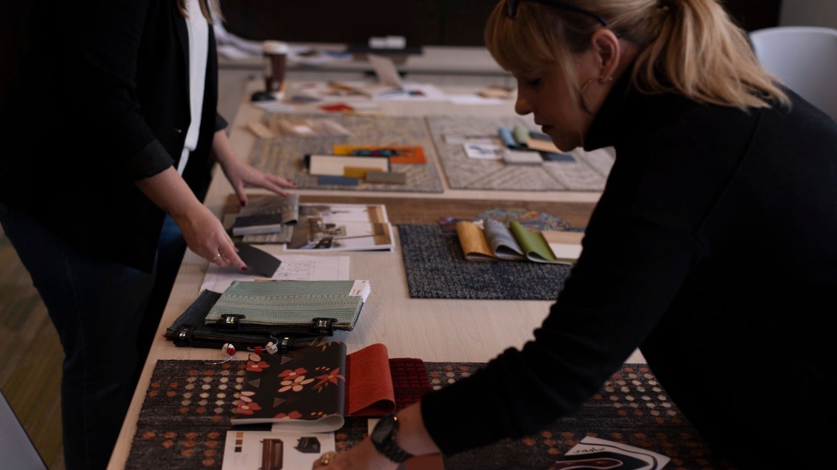 Two women looking at fabric and design samples spread out on a large table, with various fabric swatches, color samples, and catalogs in a meeting or design studio.
