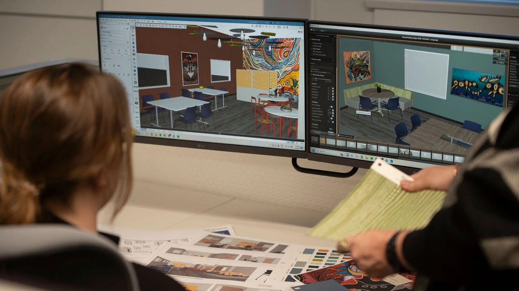 Two people working on interior design or architecture projects at a desk with dual computer monitors displaying 3D renderings of interior spaces. One person is holding printed color swatches and documents, with additional printed materials and sketch
