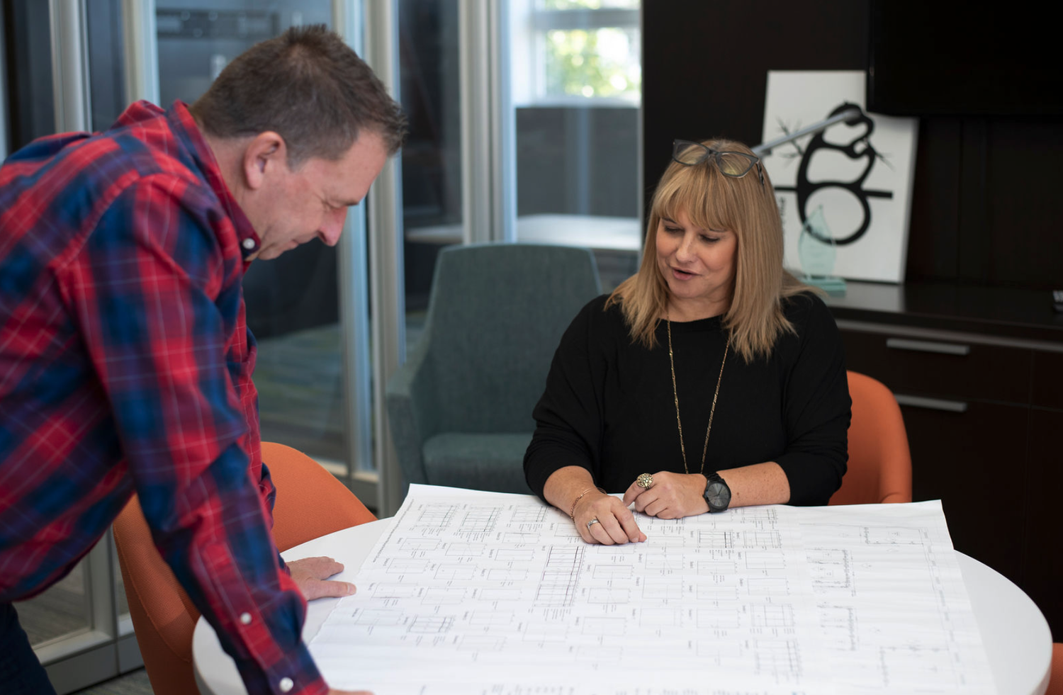 A man and woman examining large architectural blueprints on a white table in an office setting.