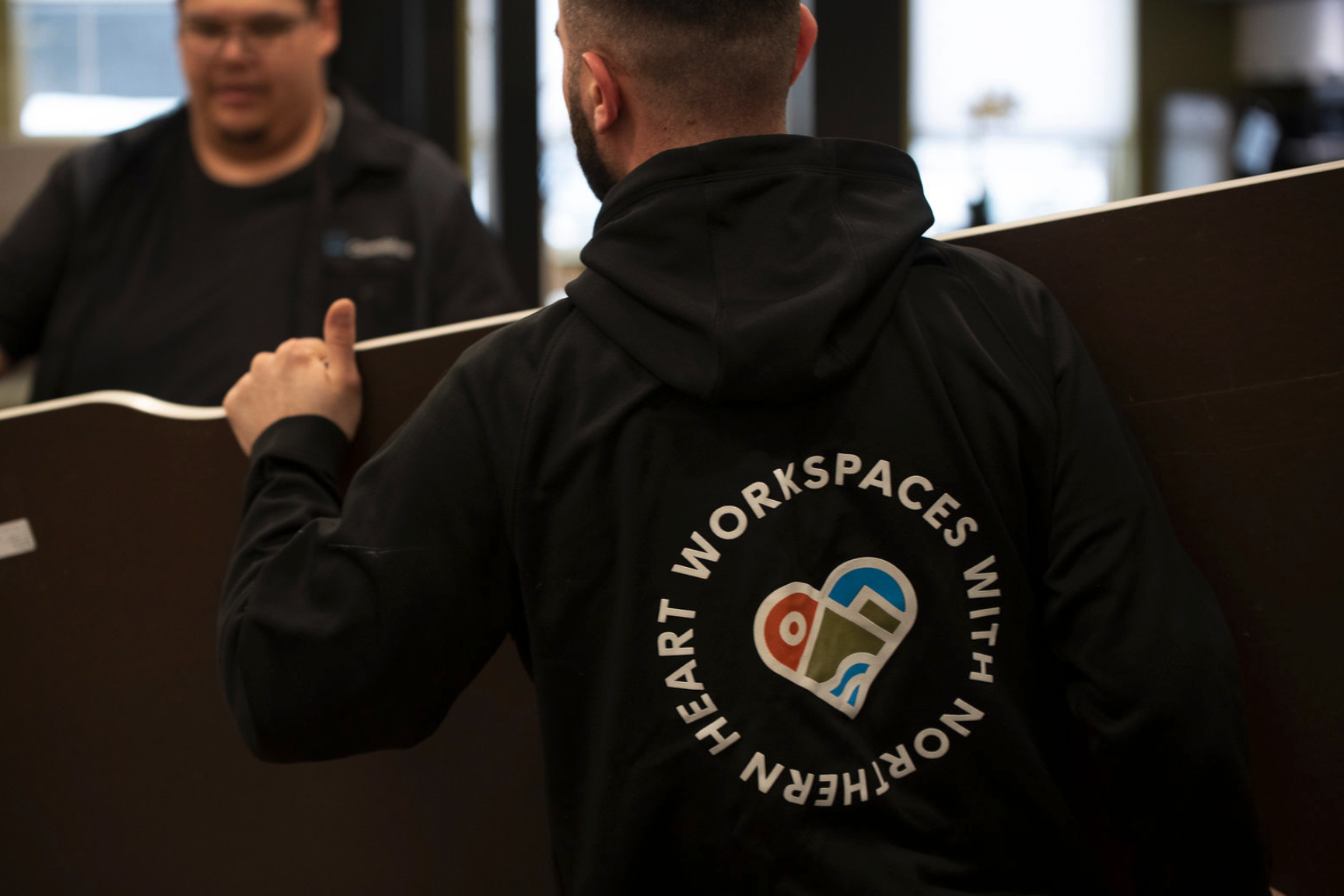 Person wearing a black jacket with 'HEART WORKSPACES WITHIN NOIRFFREN' and a heart-shaped logo on the back, holding a wooden board, in indoor setting.