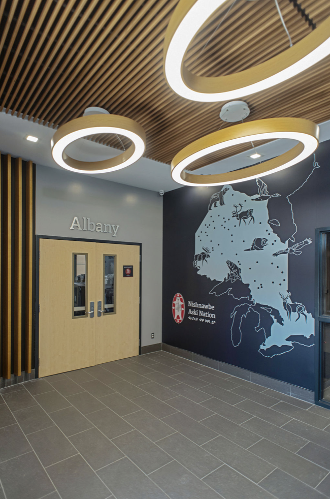 Interior view of a building with a modern ceiling design featuring circular LED light fixtures, a gray wall with the word 'Albany,' a mural of Alaska with animal illustrations, and a door marked with the Smithsonian Institution logo for the Nishnawbe Aski Nation.