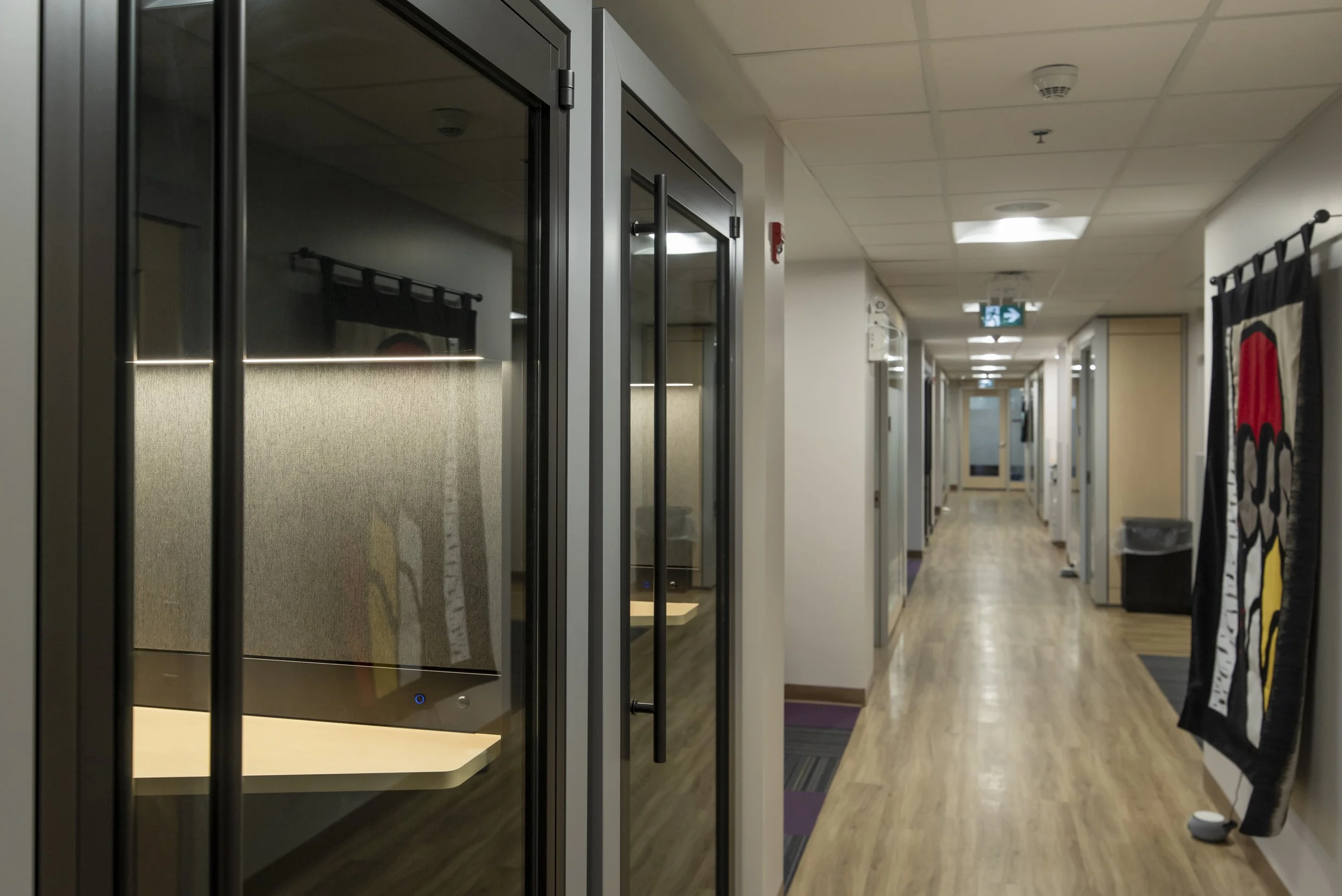 Empty office hallway with a glass-enclosed phone booth on the left side, a colorful tapestry with a stylized design on the right, and doors along the hallway leading to additional offices or rooms.