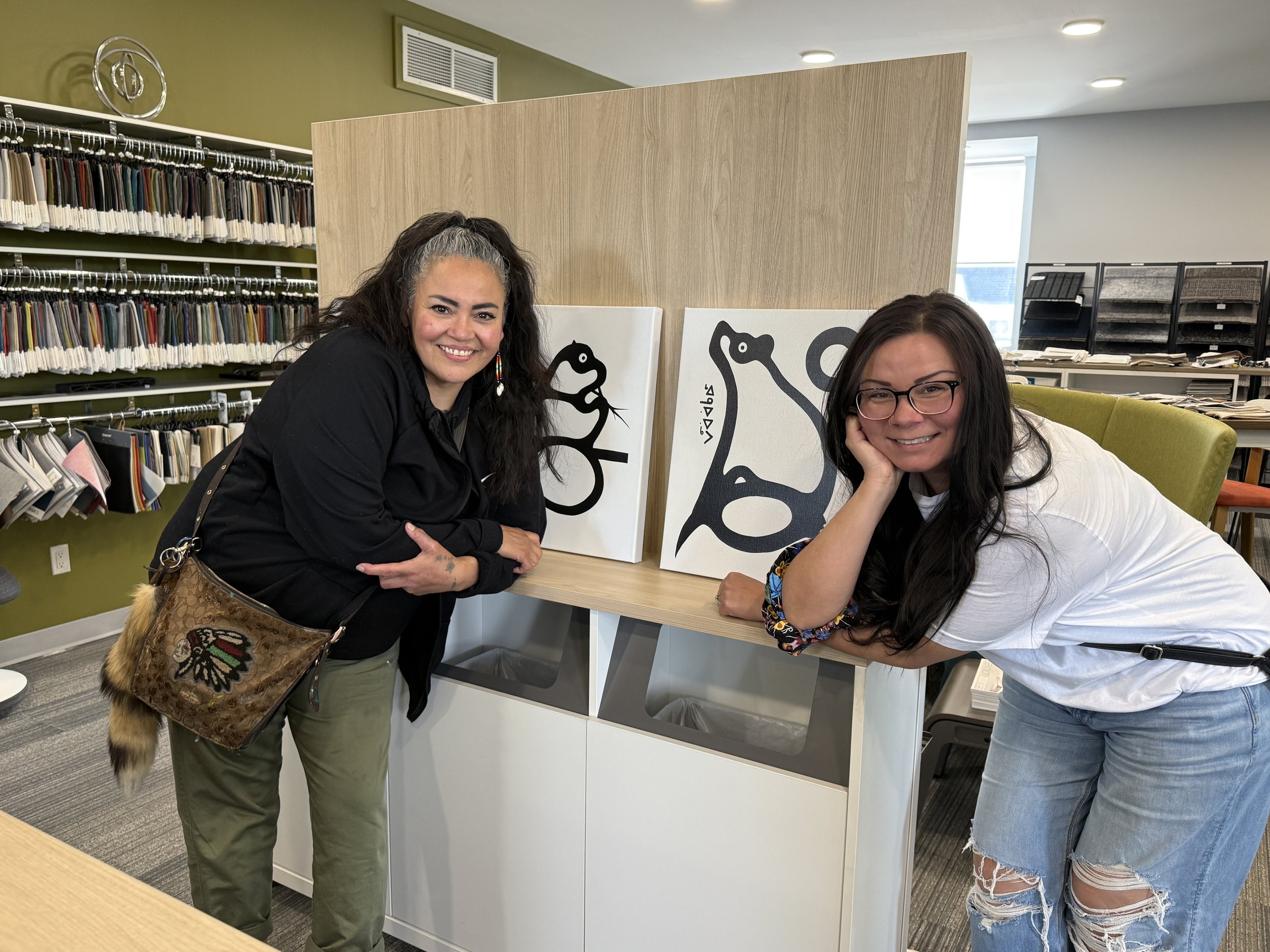 Two women smiling and posing beside abstract animal artwork in a modern interior space, with shelves of fabric samples in the background.