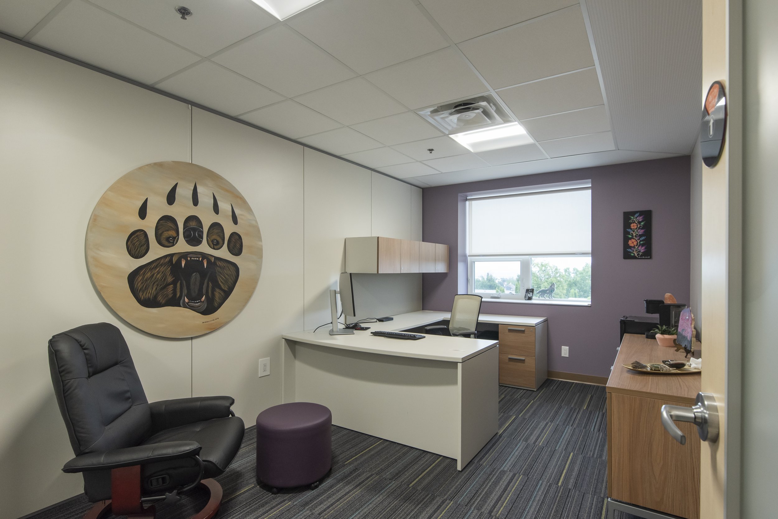 Office reception area with a bear paw wall art, a black office chair, a purple ottoman, a white desk with a computer, and a window overlooking green trees.