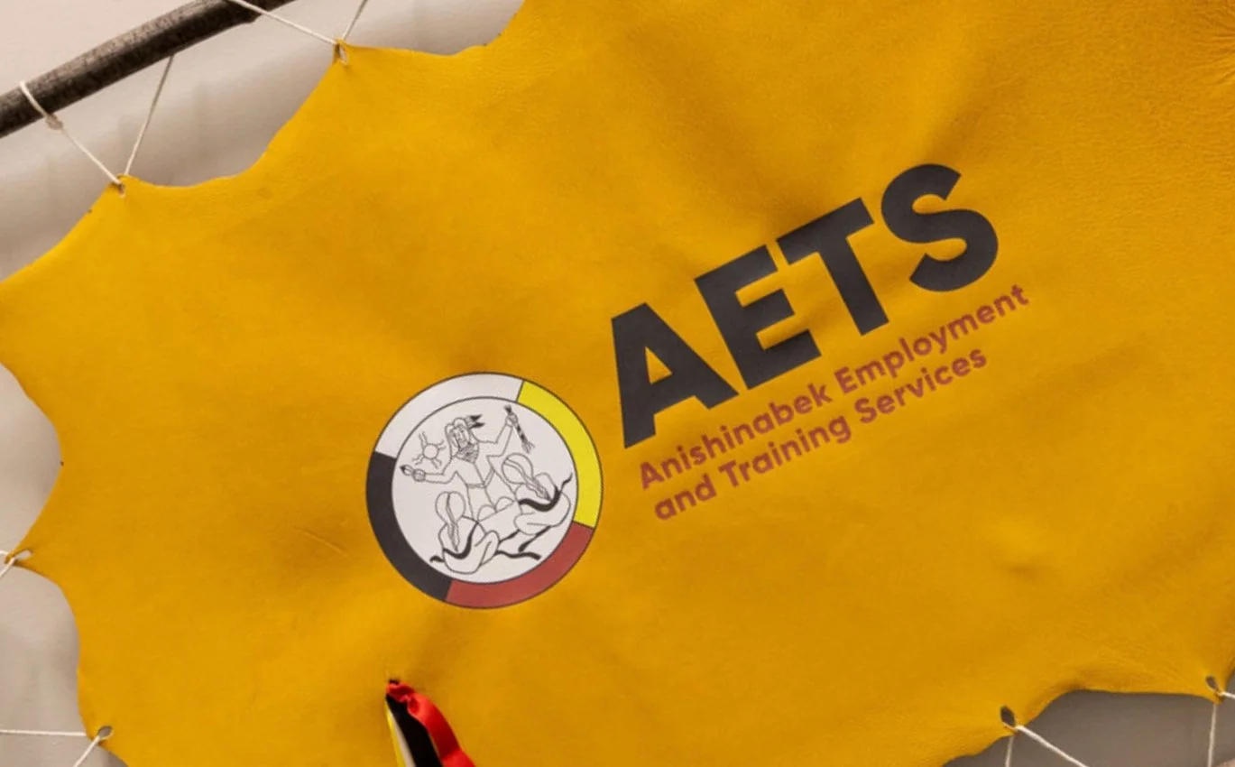 Yellow wall hanging with the logo and text for Anishinabek Employment and Training Services (AETS), featuring a circular emblem with a black, white, yellow, and red border and a line drawing of a person holding a tool.