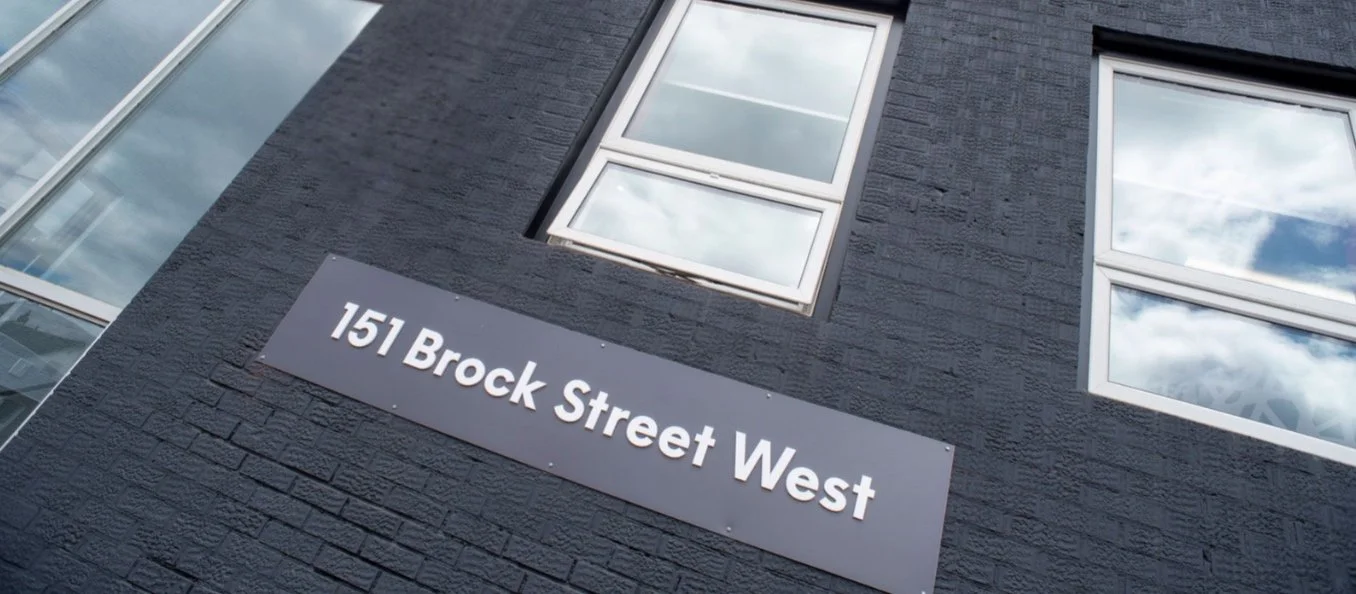 Street sign mounted on a black brick building reads '151 Brock Street West' under windows reflecting clouds in the sky.