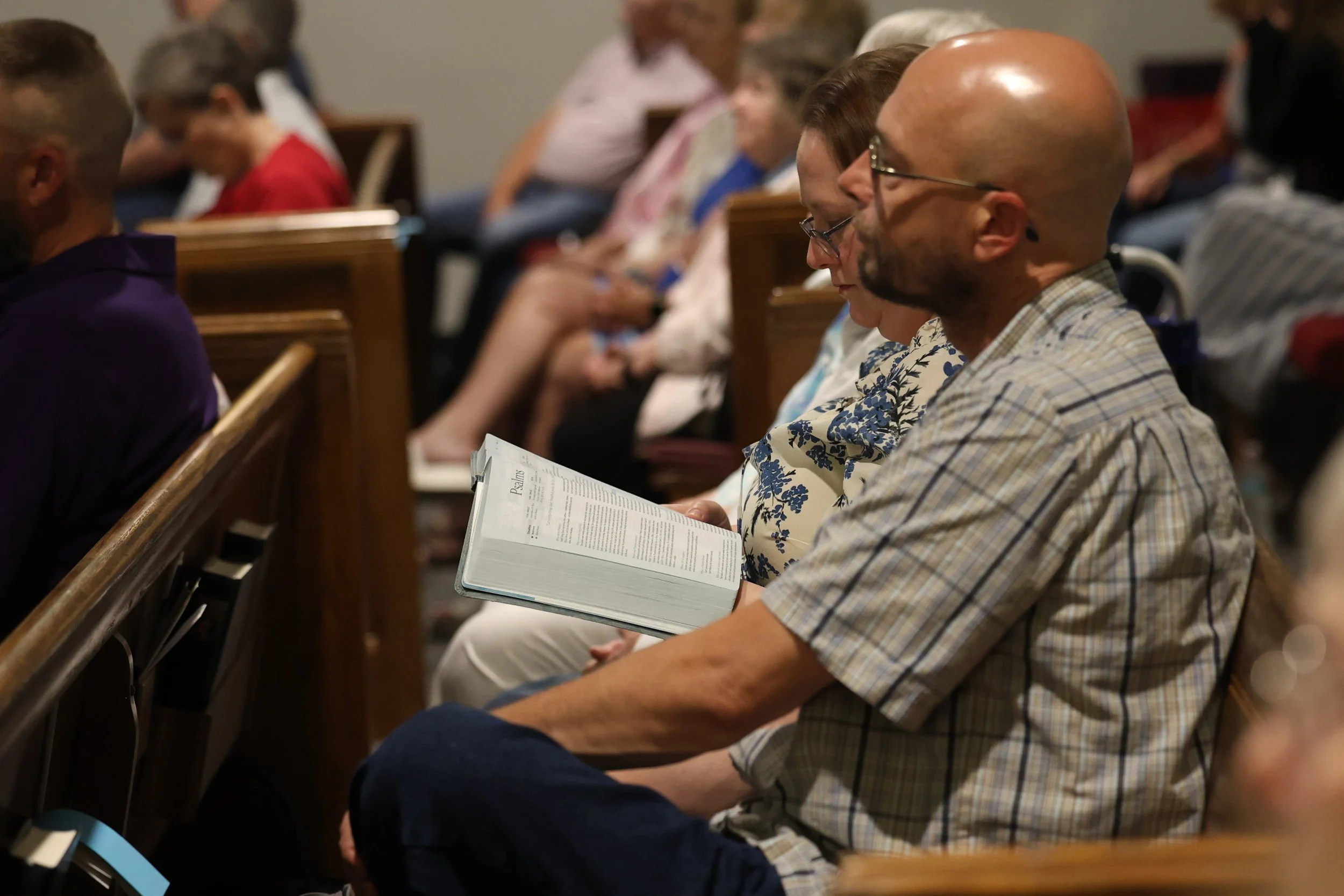 People seated in pews inside a church, engaged in prayer or reflection; some are reading hymnals or service programs.