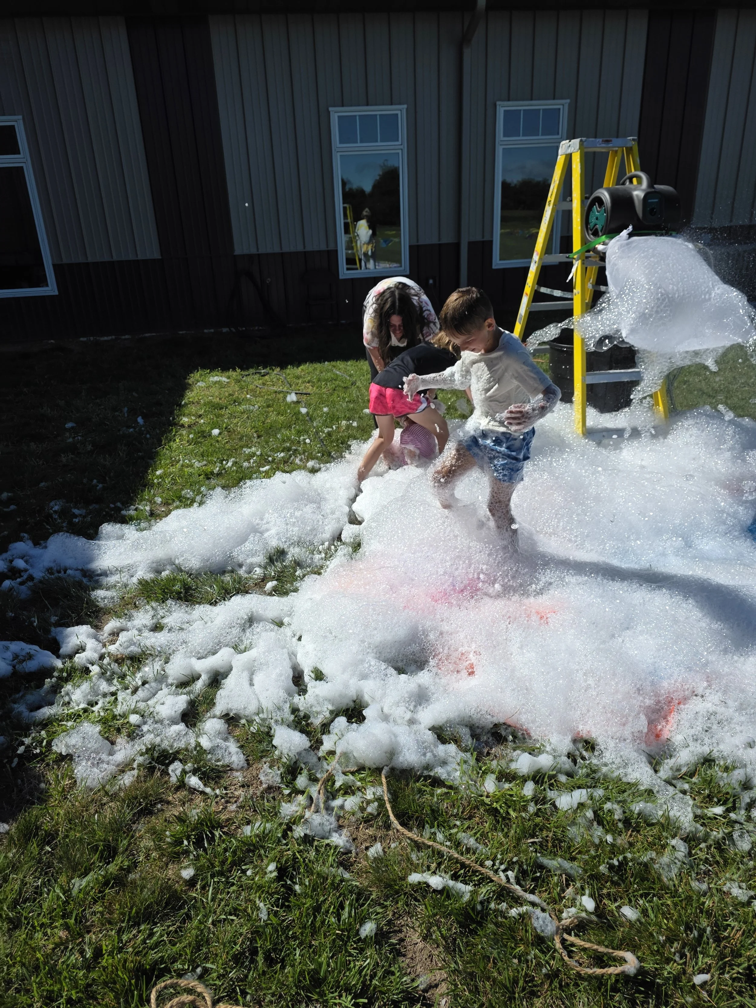 Children playing outside in foam created by a foam machine, with grass, a wooden building, and windows in the background.