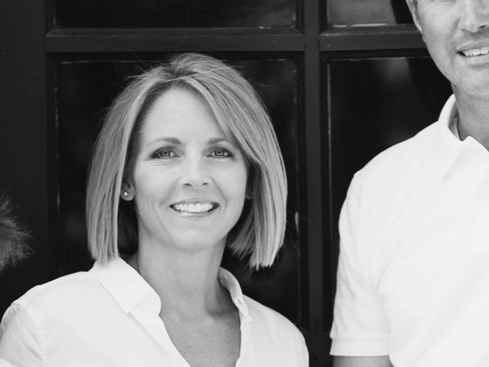 A woman with shoulder-length blonde hair wearing a white shirt, smiling in front of a dark background.