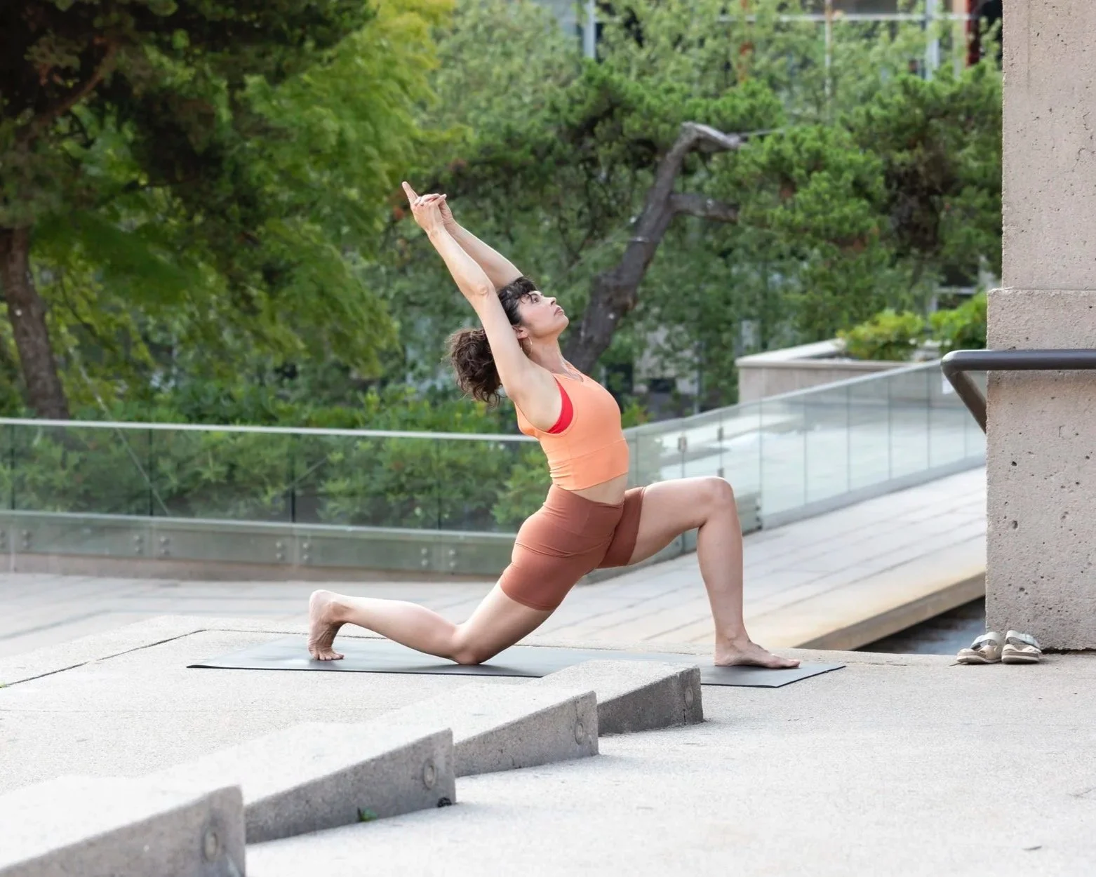 Woman doing yoga outdoors on a mat, lunging with arms raised overhead against a backdrop of trees and a glass railing.
